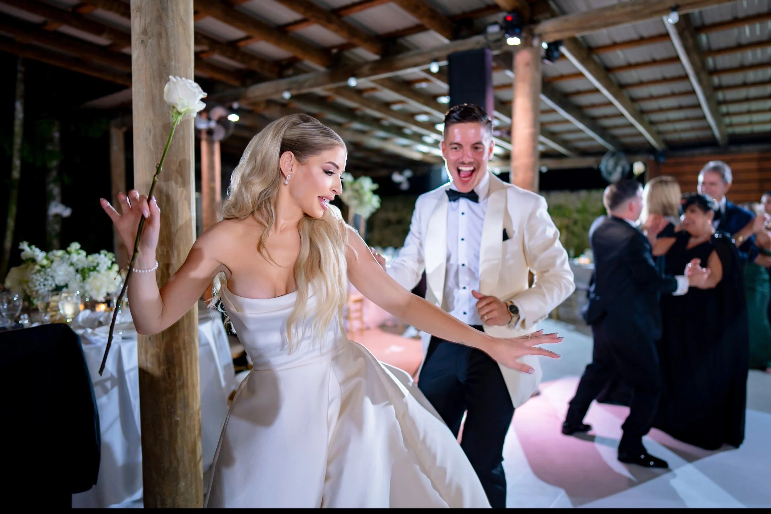 Bride and groom dancing happily at their wedding reception, with guests dancing in the background, under a wooden pavilion with floral decorations.
