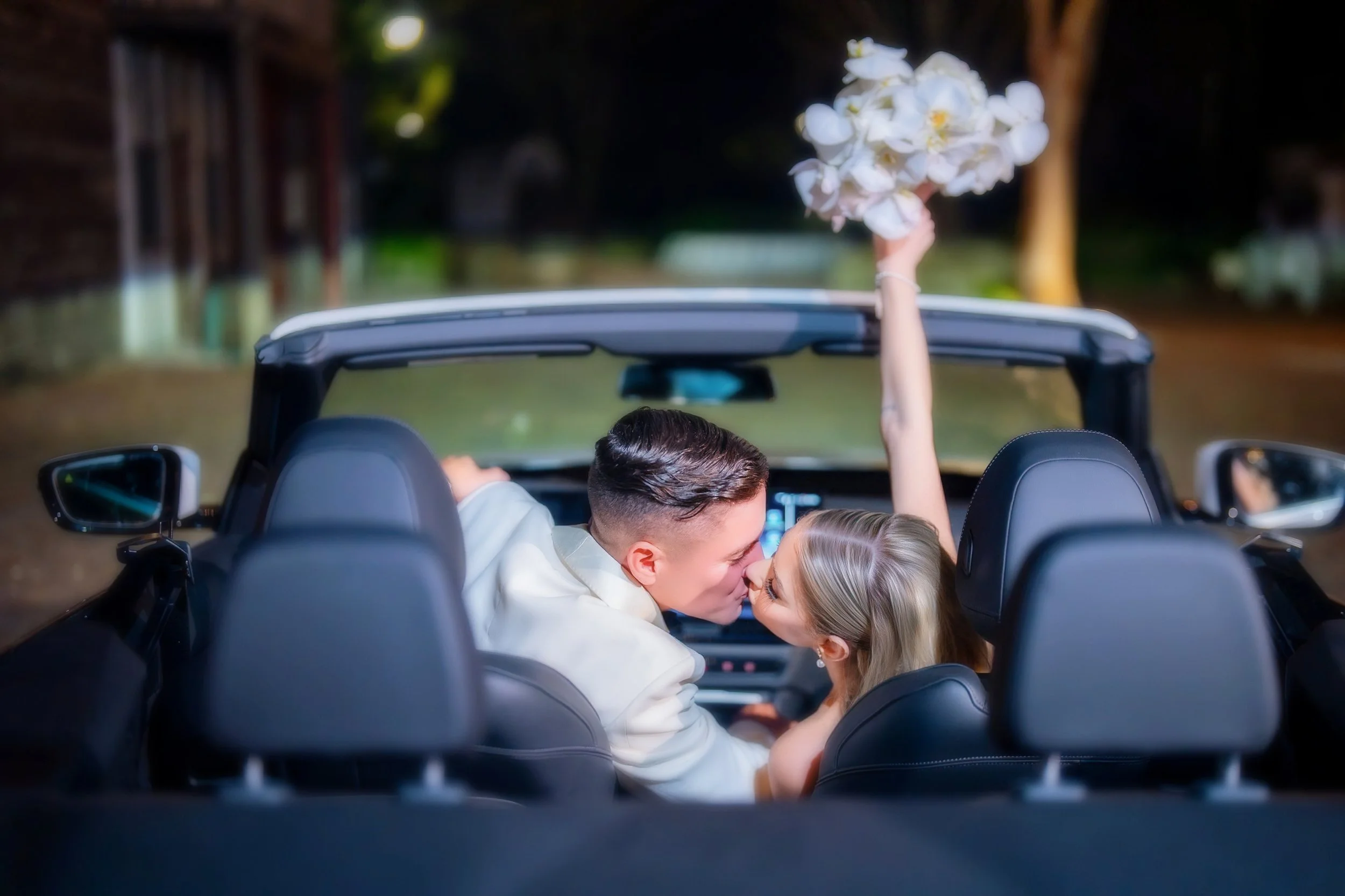 A couple in a convertible car sharing a kiss, with the woman holding a bouquet of white flowers in the air, during what appears to be their wedding or special celebration.