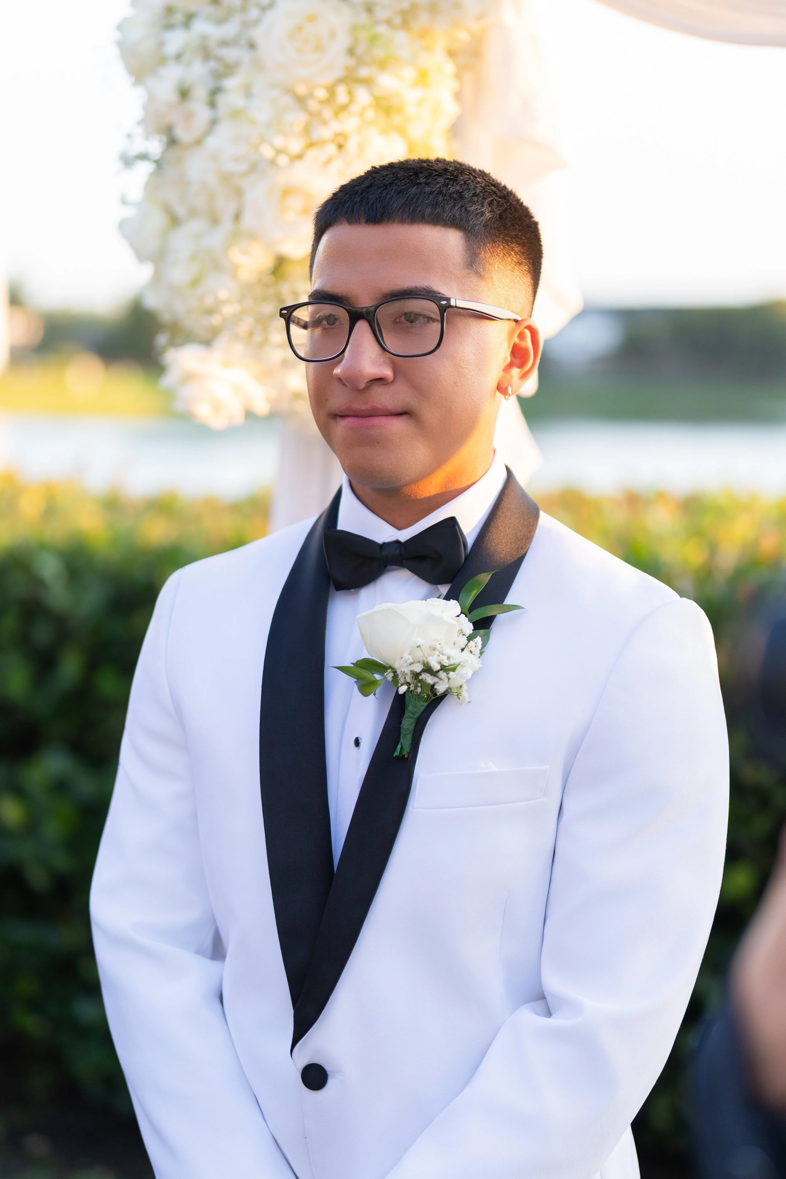 A young man in a white tuxedo with a black lapel and black bowtie, wearing glasses and a boutonniere, standing outdoors during a wedding ceremony with a floral arch and a river in the background.