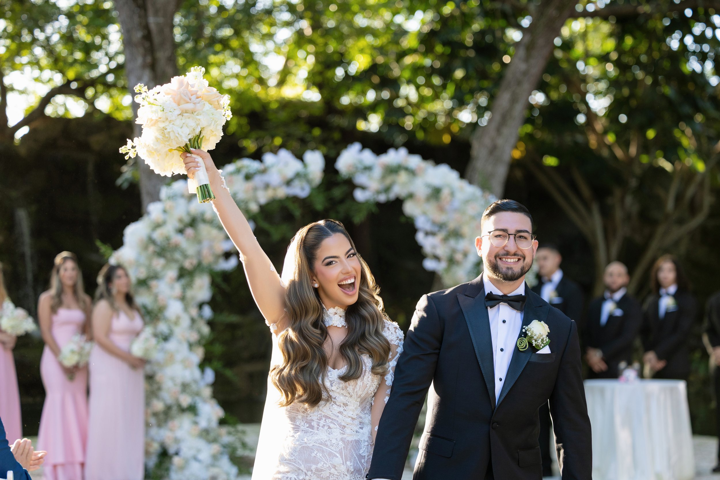Bride happily celebrating with bouquet raised in the air after wedding ceremony outside, groom beside her, with bridesmaids and groomsmen in the background.