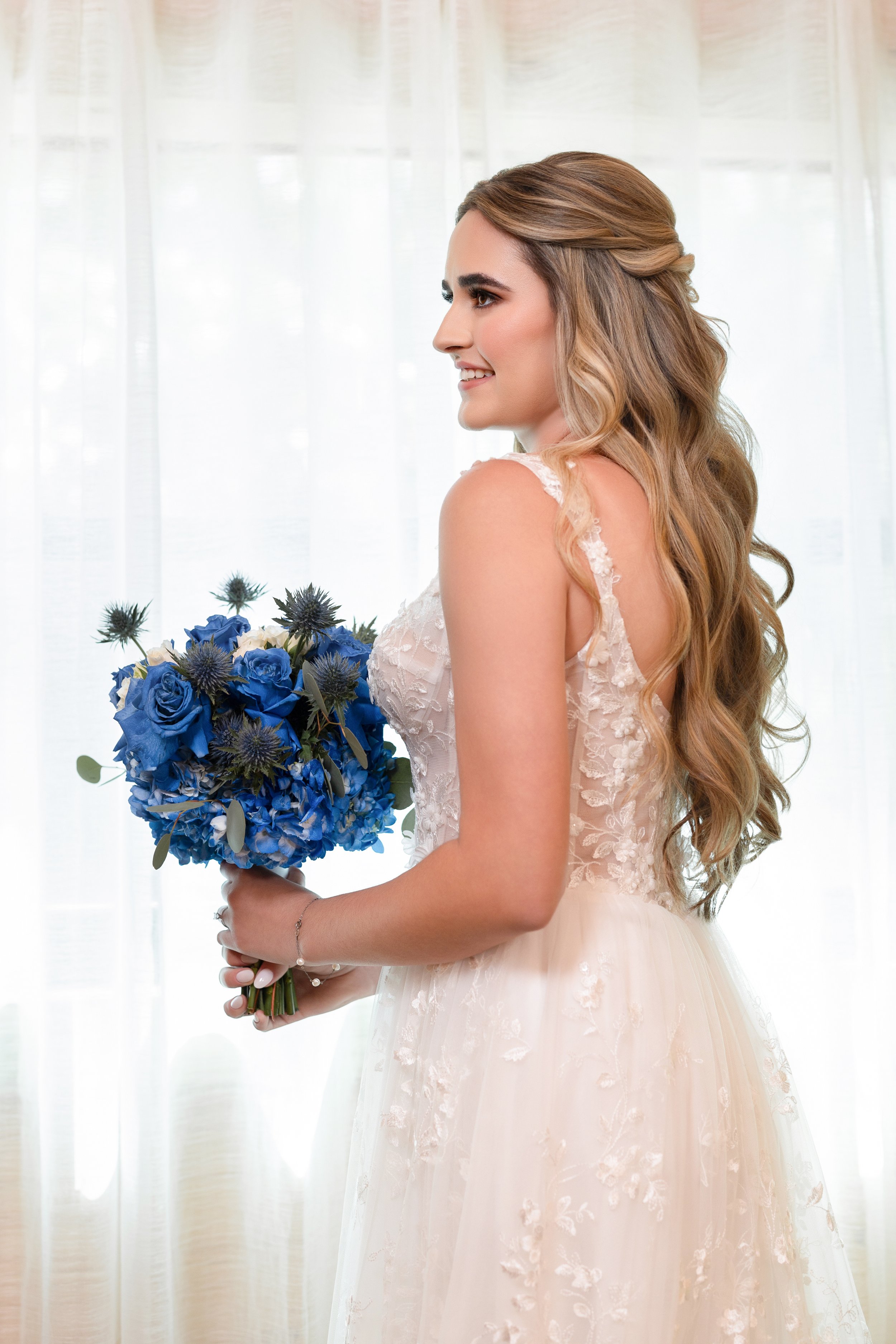 A bride with long wavy hair in a lace and tulle wedding dress holding a bouquet of blue flowers in front of a sheer white curtain.
