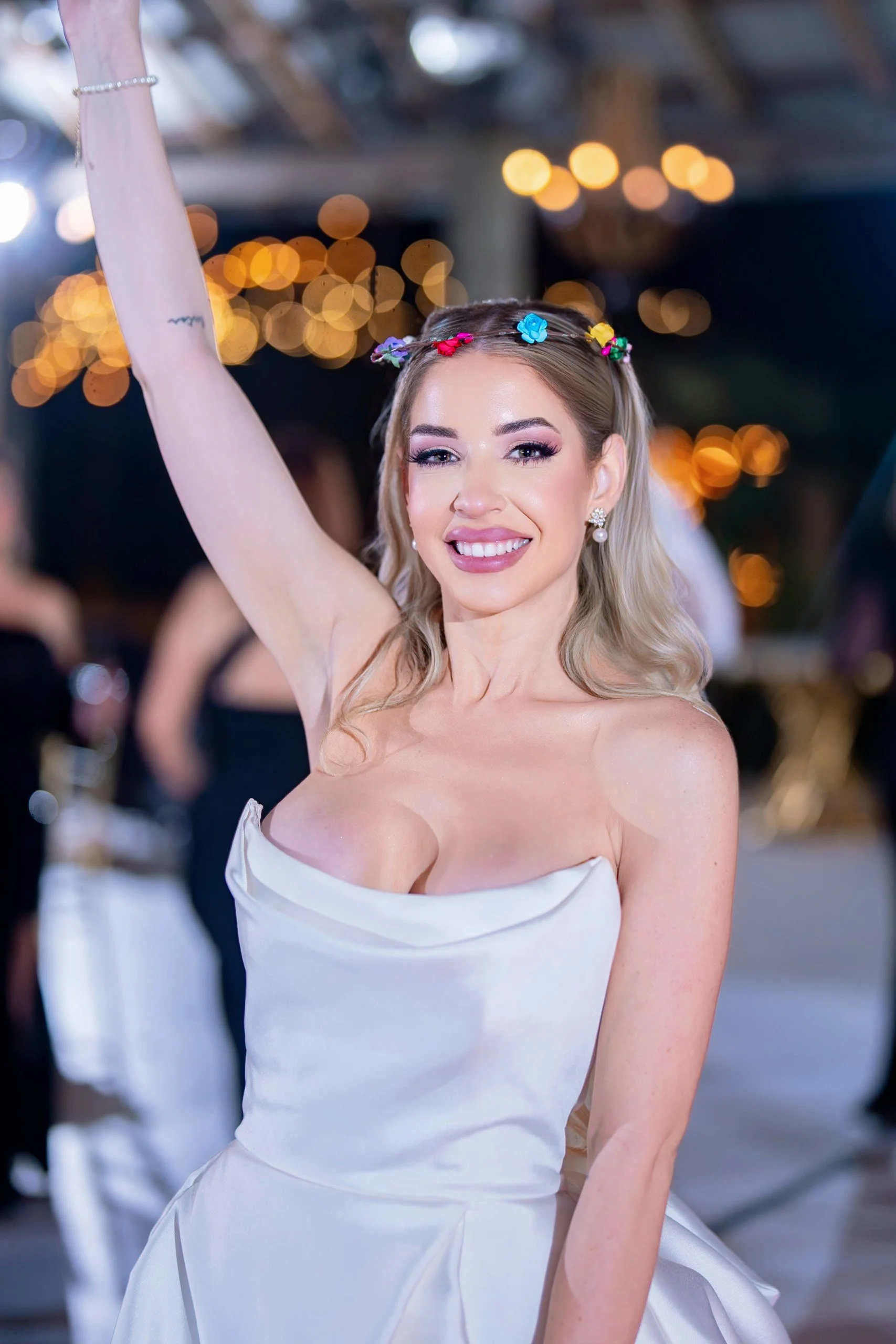 A woman smiling and dancing at a celebration or party, wearing a white dress and a colorful flower headband.