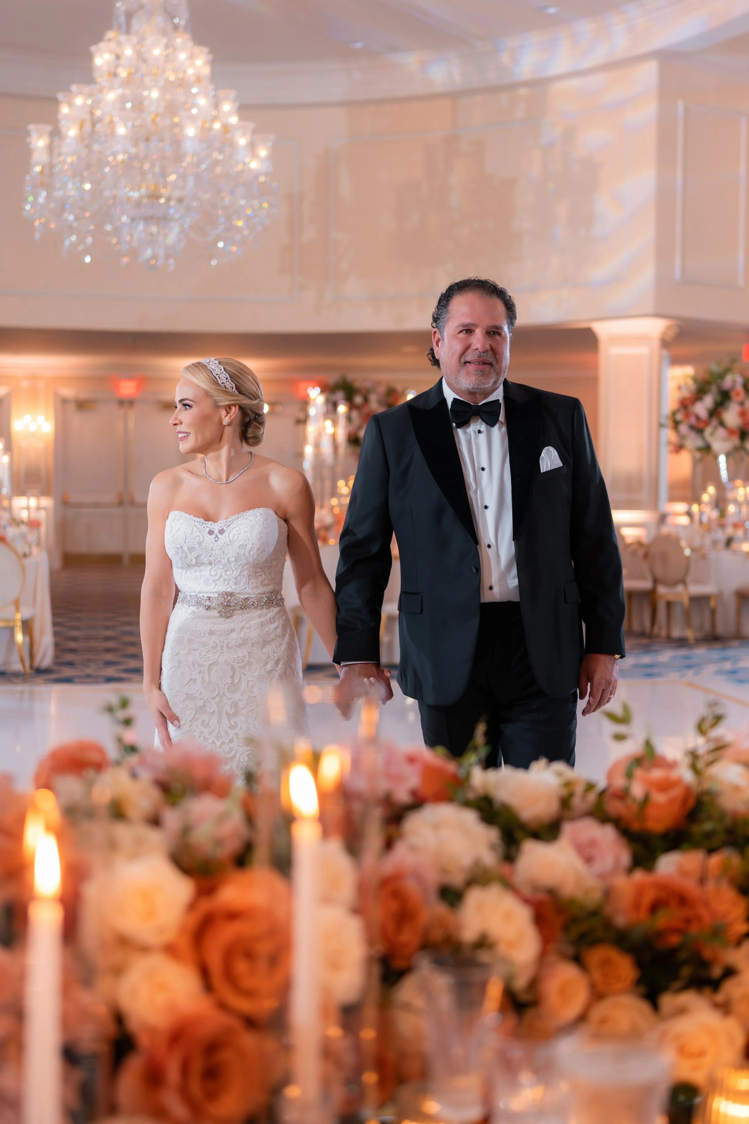 A bride and groom walk hand in hand in an elegant wedding reception hall decorated with a large chandelier, floral arrangements, candles, and luxurious decor.