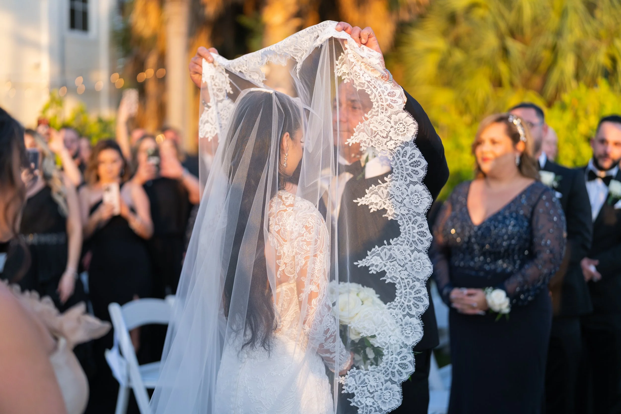 A bride and groom share a moment during their wedding ceremony outdoors, with the bride wearing a lace wedding gown and veil, and the groom lifting her veil. Guests in formal attire watch and take photos in the background.