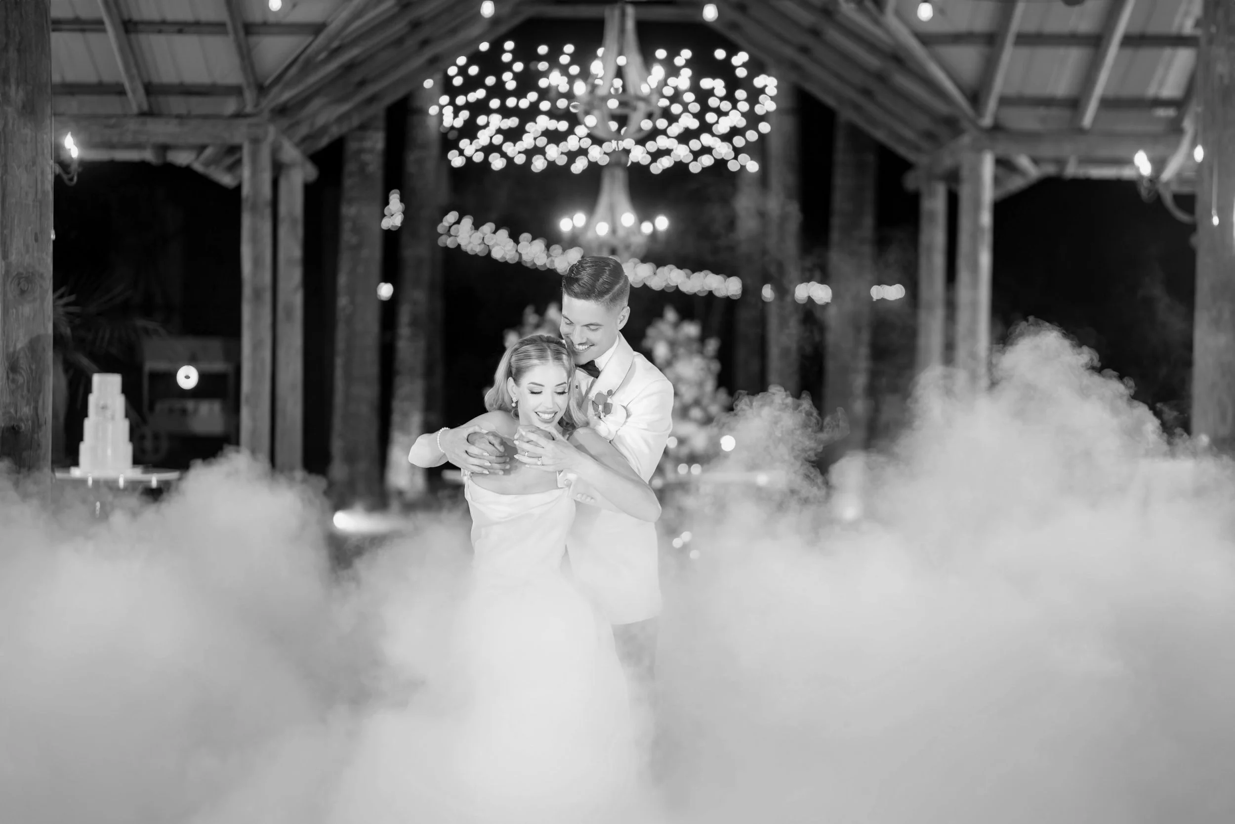 Black and white photo of a bride and groom dancing closely in a rustic barn decorated for a wedding reception with string lights and a chandelier, surrounded by fog.