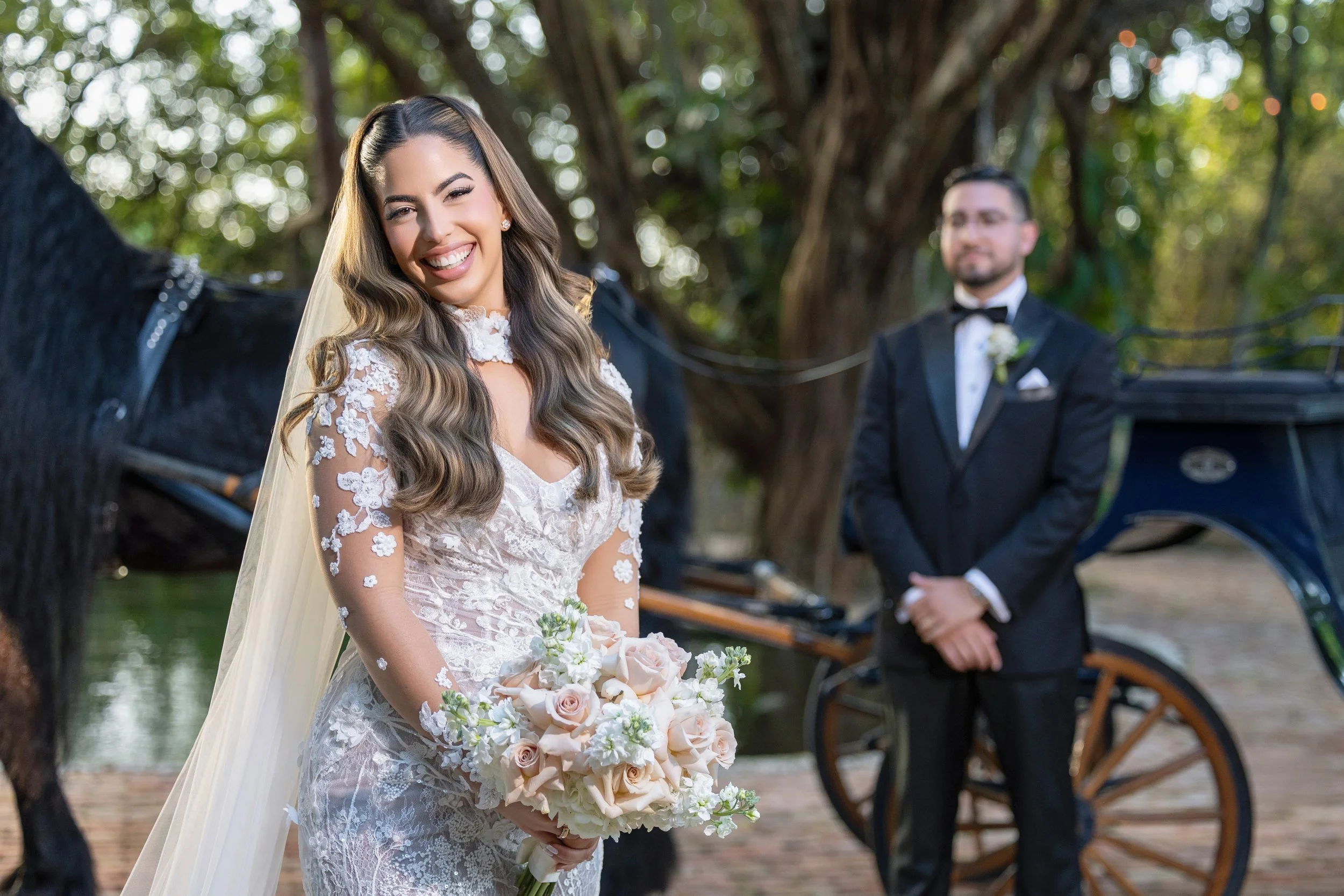 A smiling bride in a wedding dress holding a bouquet of beige and white roses, standing outdoors with a horse and a groom in a black tuxedo and bow tie in the background near a carriage.