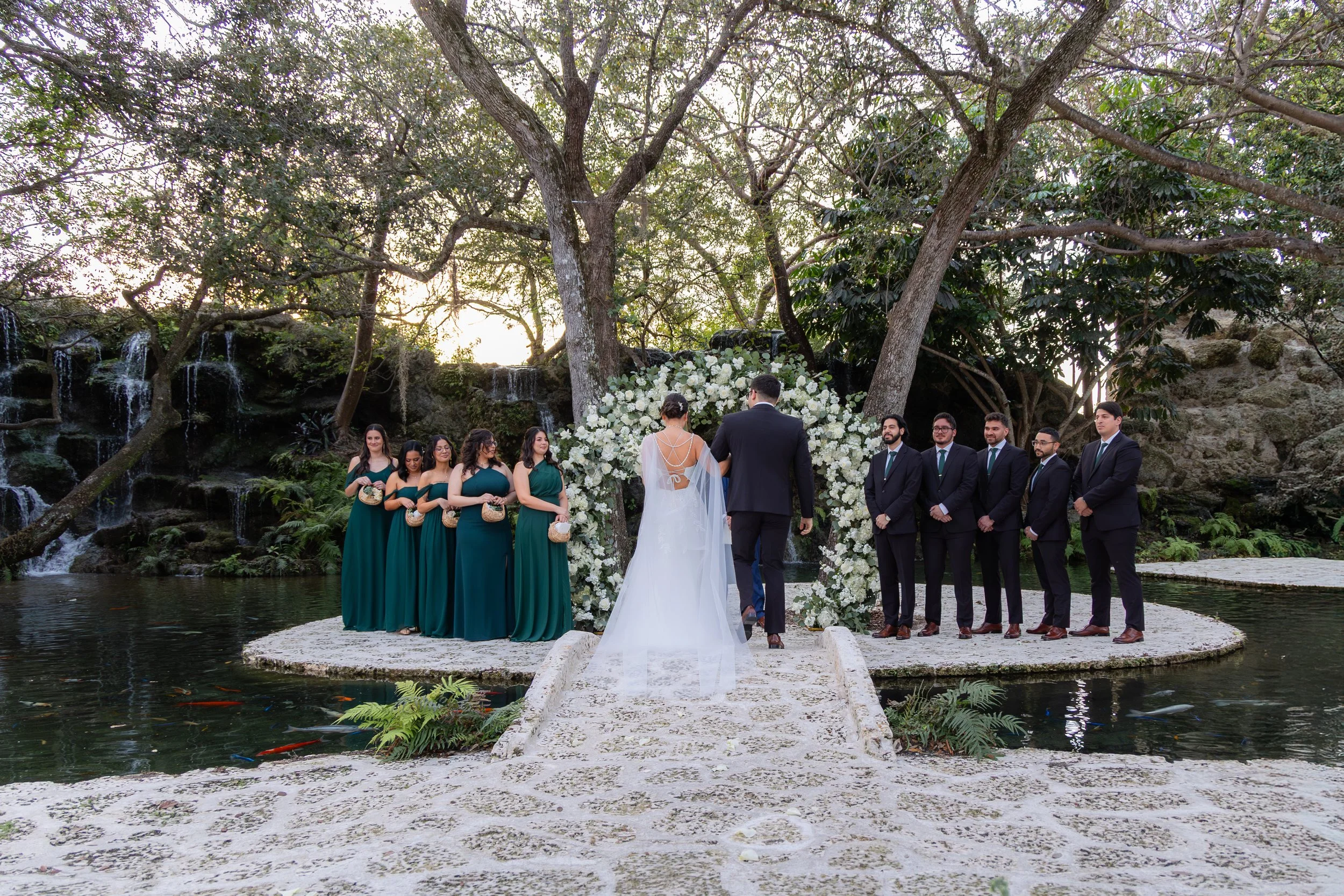A wedding ceremony taking place outdoors by a pond with a waterfall in the background, decorated with a floral arch, with the bride and groom standing at the altar and bridal party on either side.
