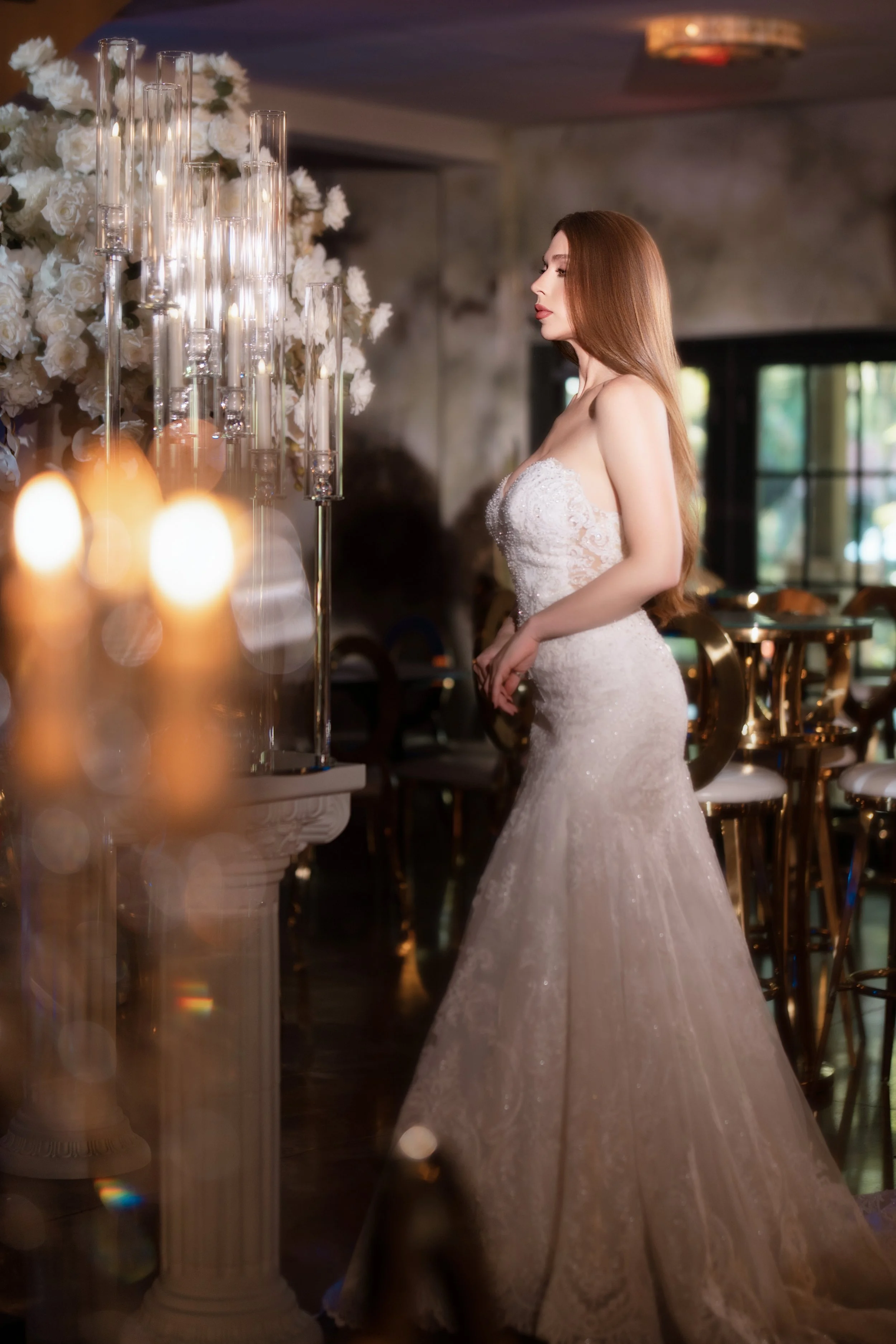 A woman in a white wedding dress standing inside a decorated venue with floral arrangements and tall candles.