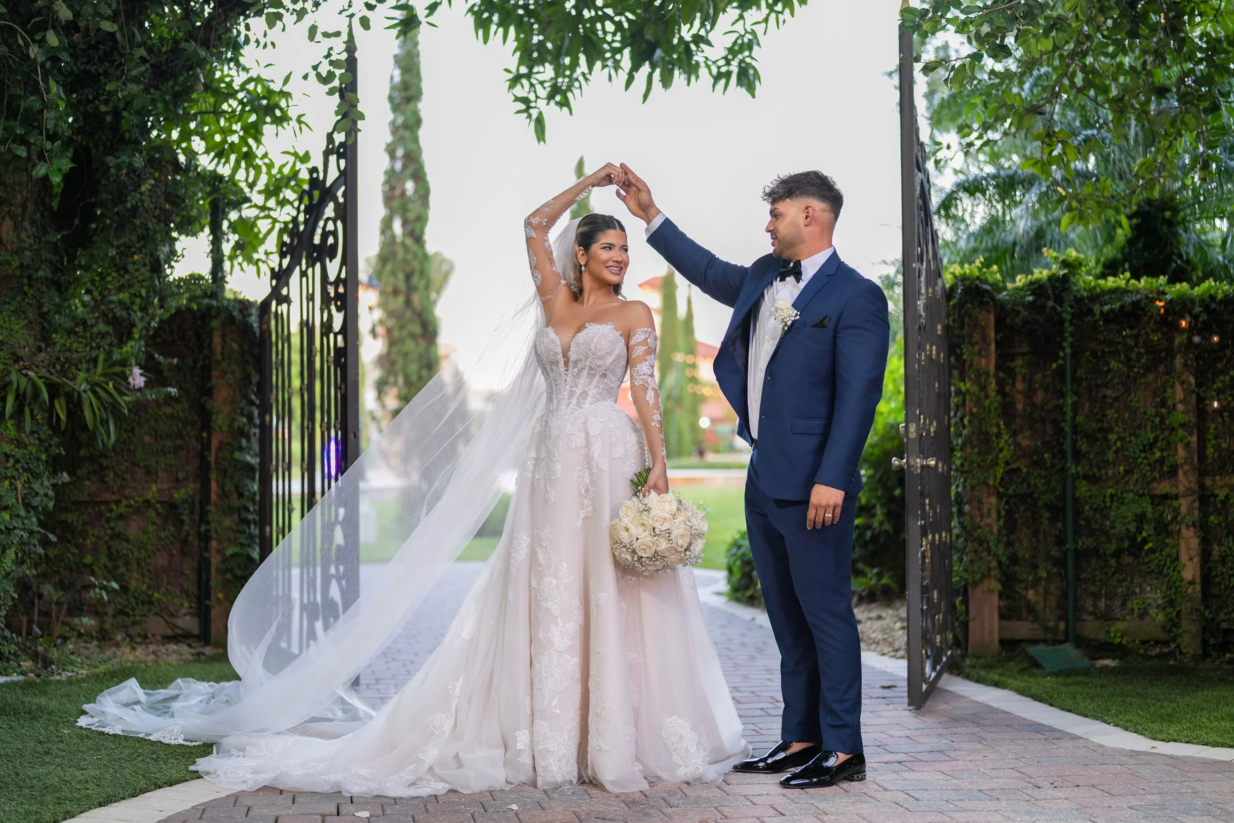 A bride and groom dancing at their wedding outdoors, with the bride in a white wedding gown holding a bouquet of white roses, and the groom in a dark blue suit with a black bow tie, standing on a brick path with open gates and greenery around.