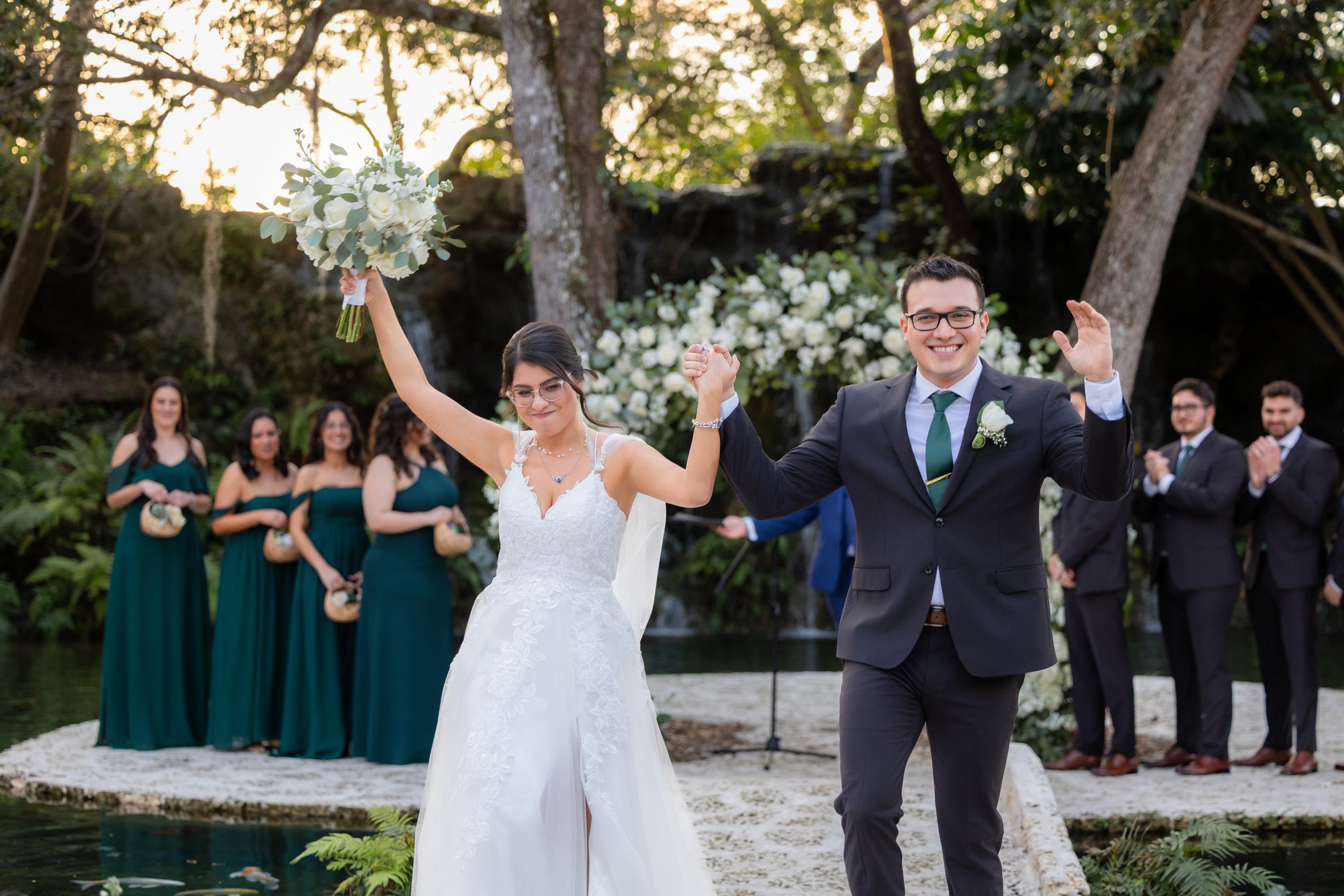 A newly married couple is celebrating their wedding outdoors by a pond, holding hands and smiling, with bridesmaids and groomsmen clapping in the background.