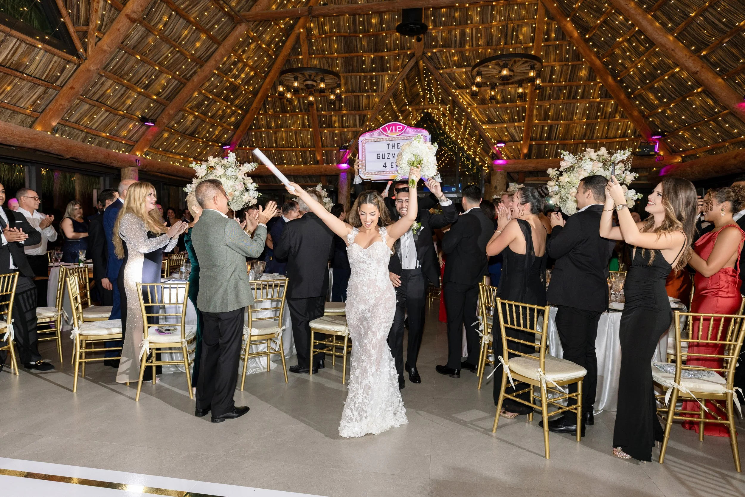 Bride in a white lace wedding dress holding a bouquet and celebrating with guests at her wedding reception in a decorated hall with string lights and floral arrangements.