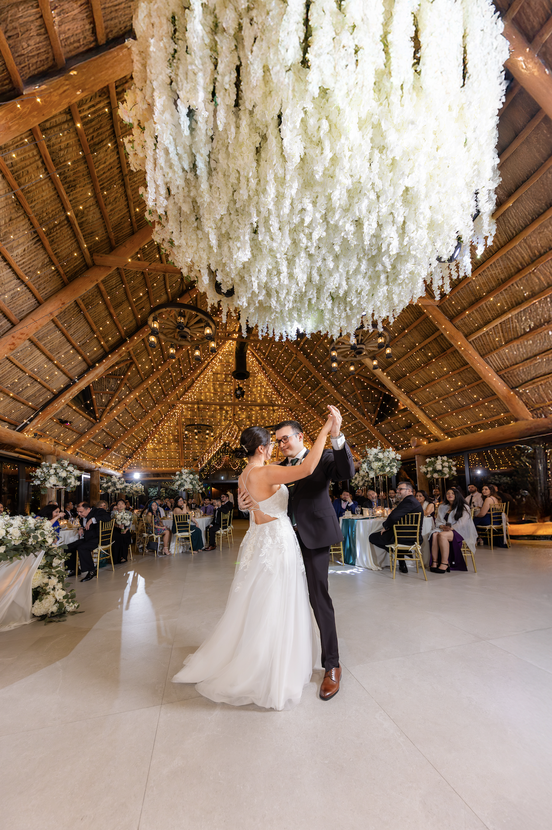 A bride and groom dance in a rustic, warmly lit wedding reception hall with hanging white floral arrangements and string lights overhead.