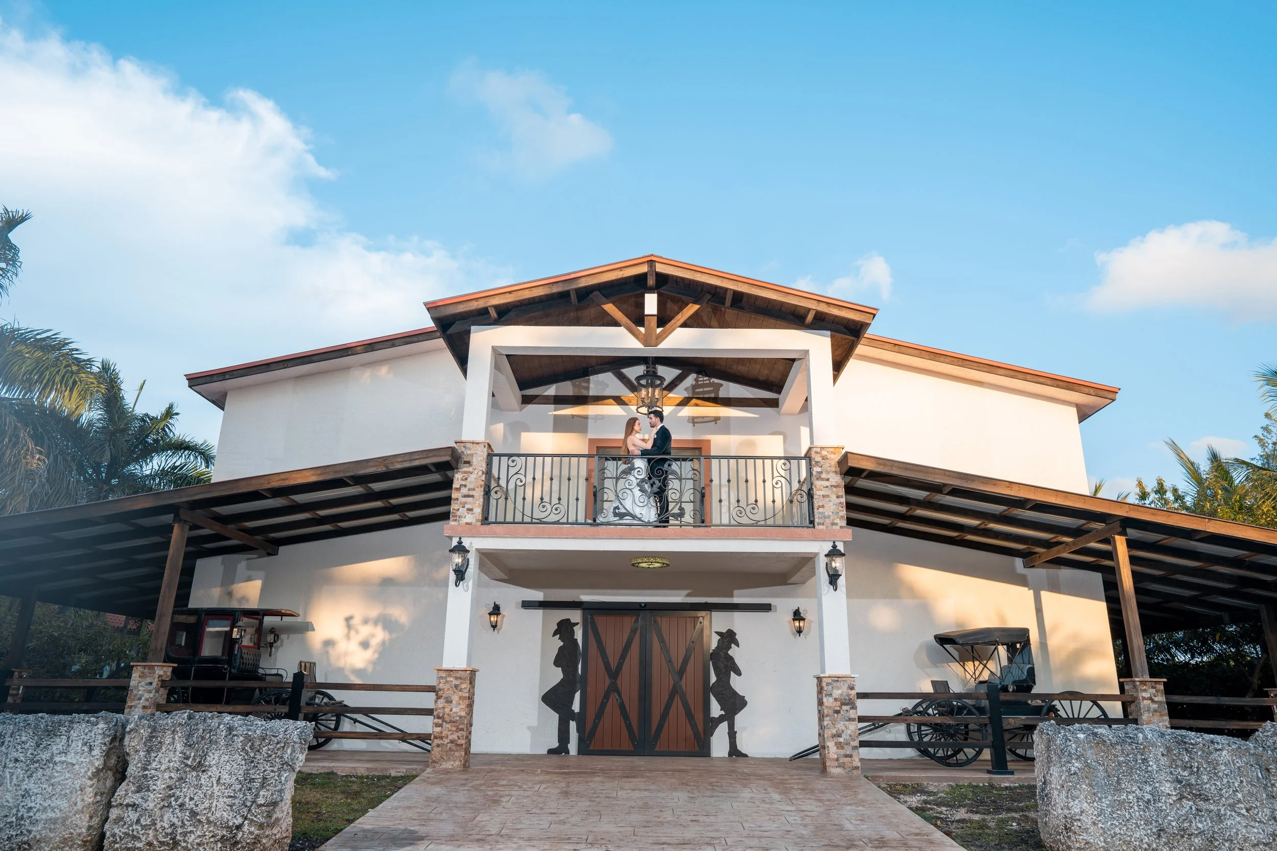 A couple getting married on a balcony of a white building with a wooden roof and decorative wrought iron railings, overlooking a paved courtyard with statues of dancing women and old-fashioned horse-drawn carriages on either side.