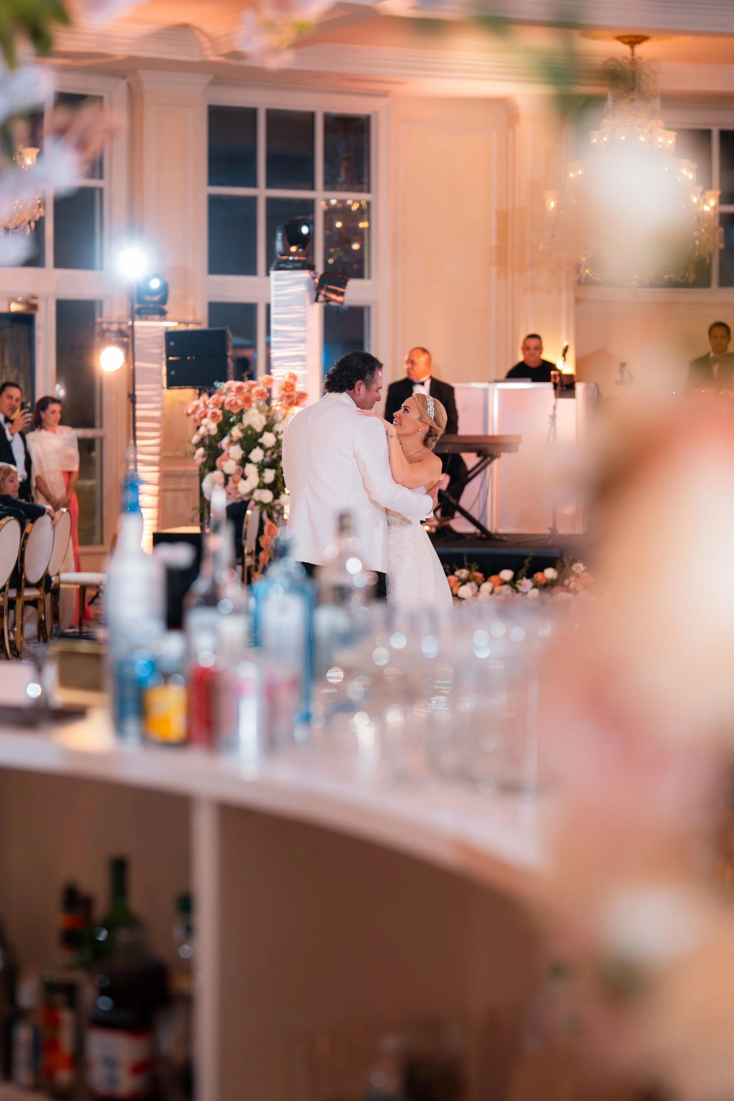 A bride and groom share their first dance at a wedding reception with a decorated floral backdrop, guests, and a music band in the background.