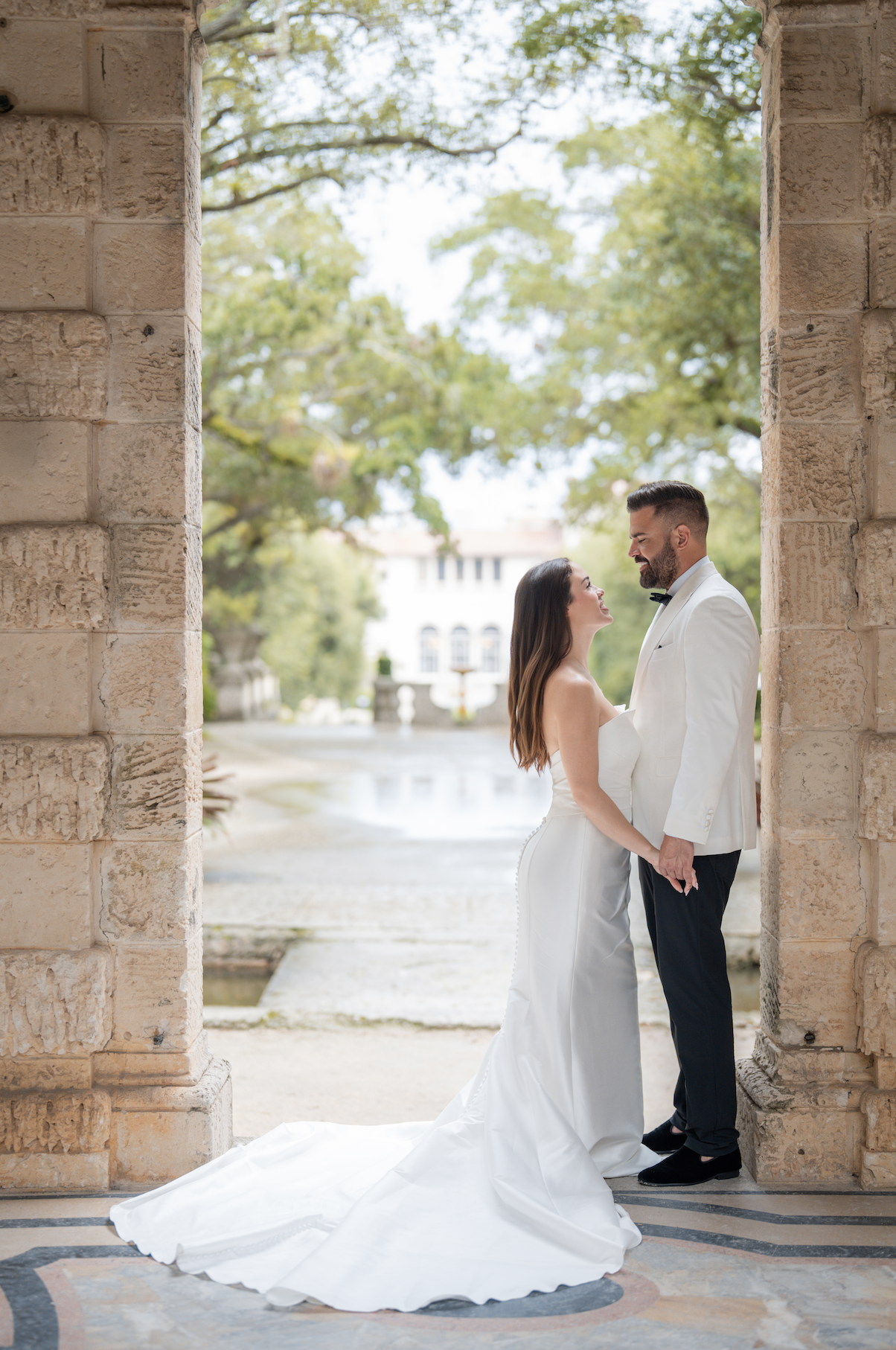A bride and groom holding hands and looking into each other's eyes outside, framed by a stone archway with trees and a building in the background.