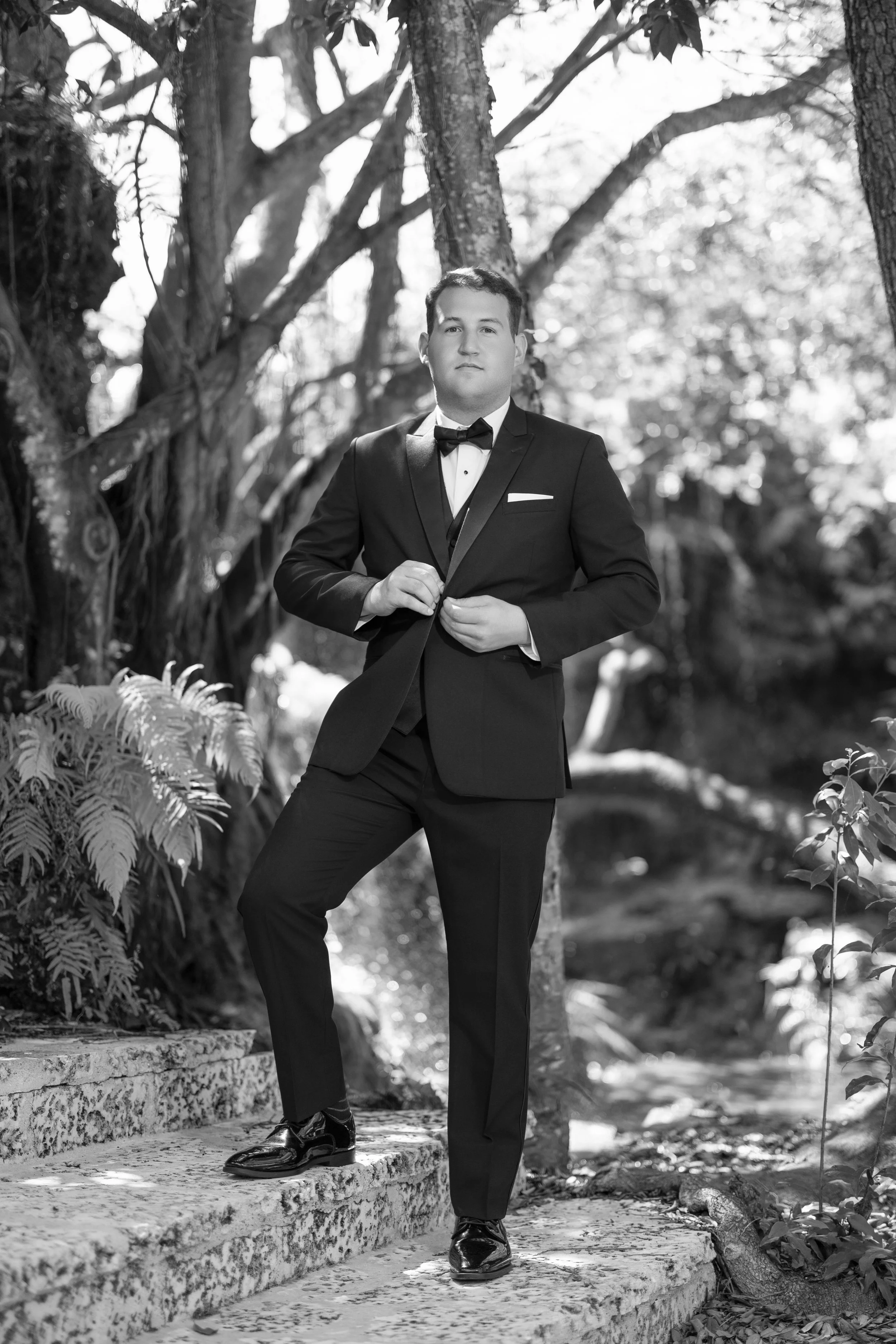 A man in a tuxedo with a bowtie, standing outdoors on stone steps in a forested area, adjusting his jacket.