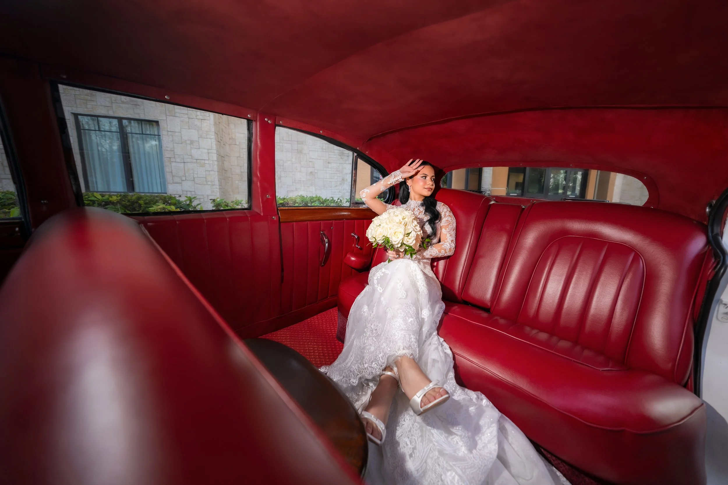 Bride sitting inside a vintage red car, wearing a white wedding dress and holding a bouquet of white roses.