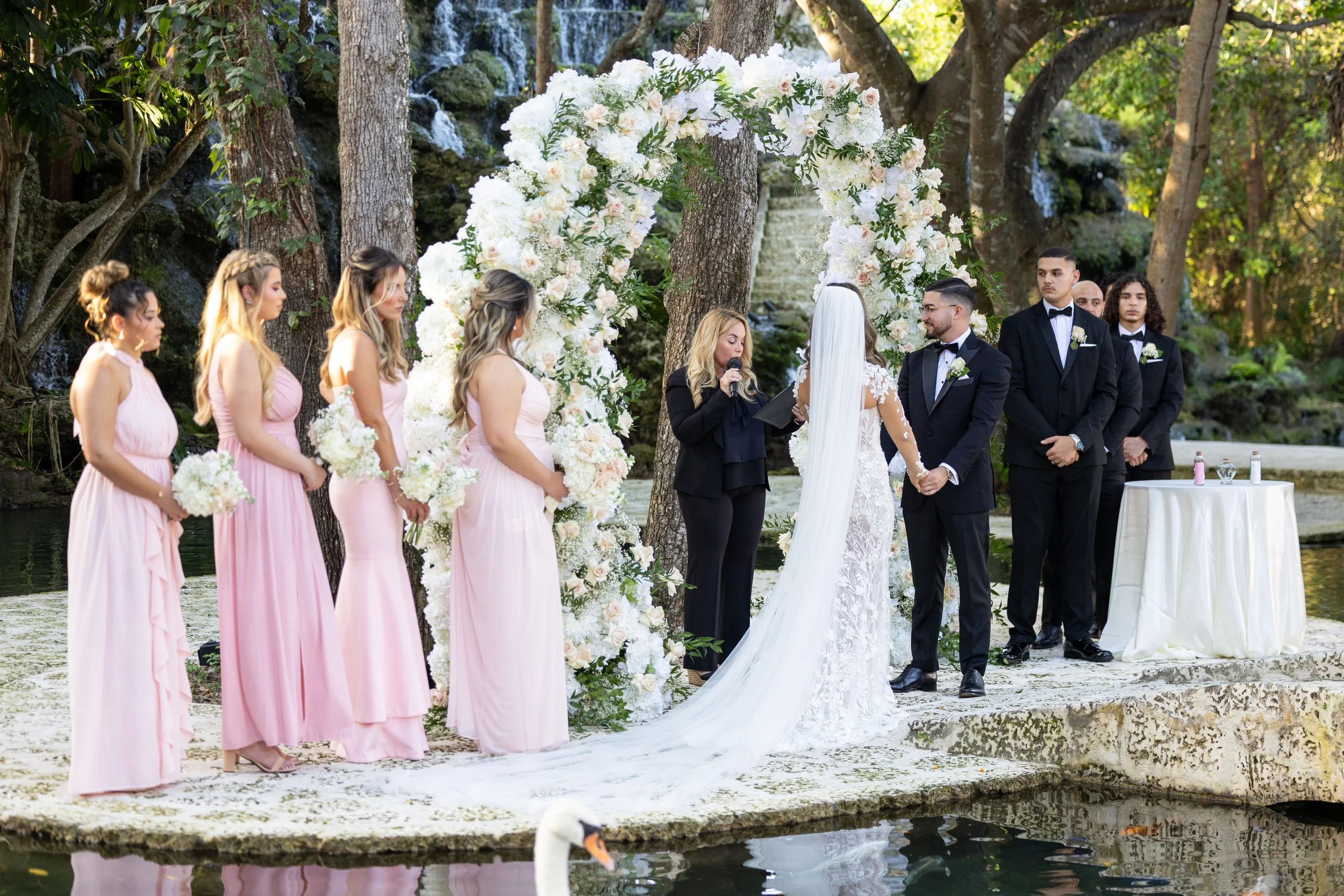 Wedding ceremony with bride and groom holding hands, surrounded by bridesmaids in pink dresses and groomsmen in black suits outdoors, with a floral arch, on a stone platform by water with a swan.