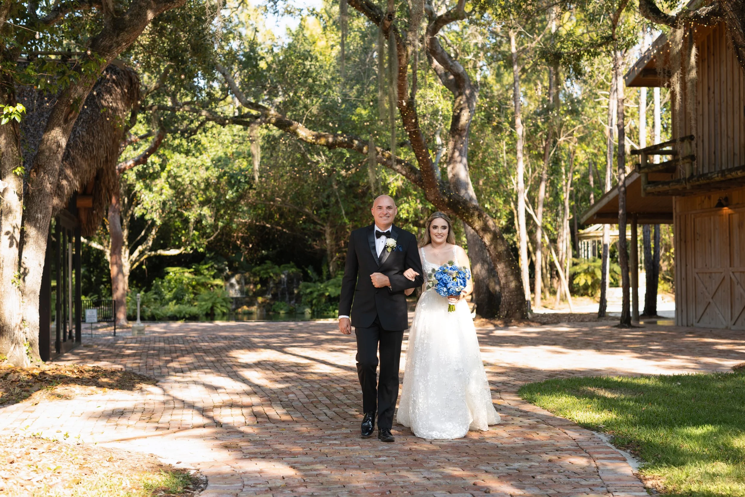 Bride in a white wedding dress holding a bouquet of blue flowers walking arm-in-arm with the groom in a black tuxedo and bow tie, on a brick path in a wooded outdoor setting with trees and wooden buildings.