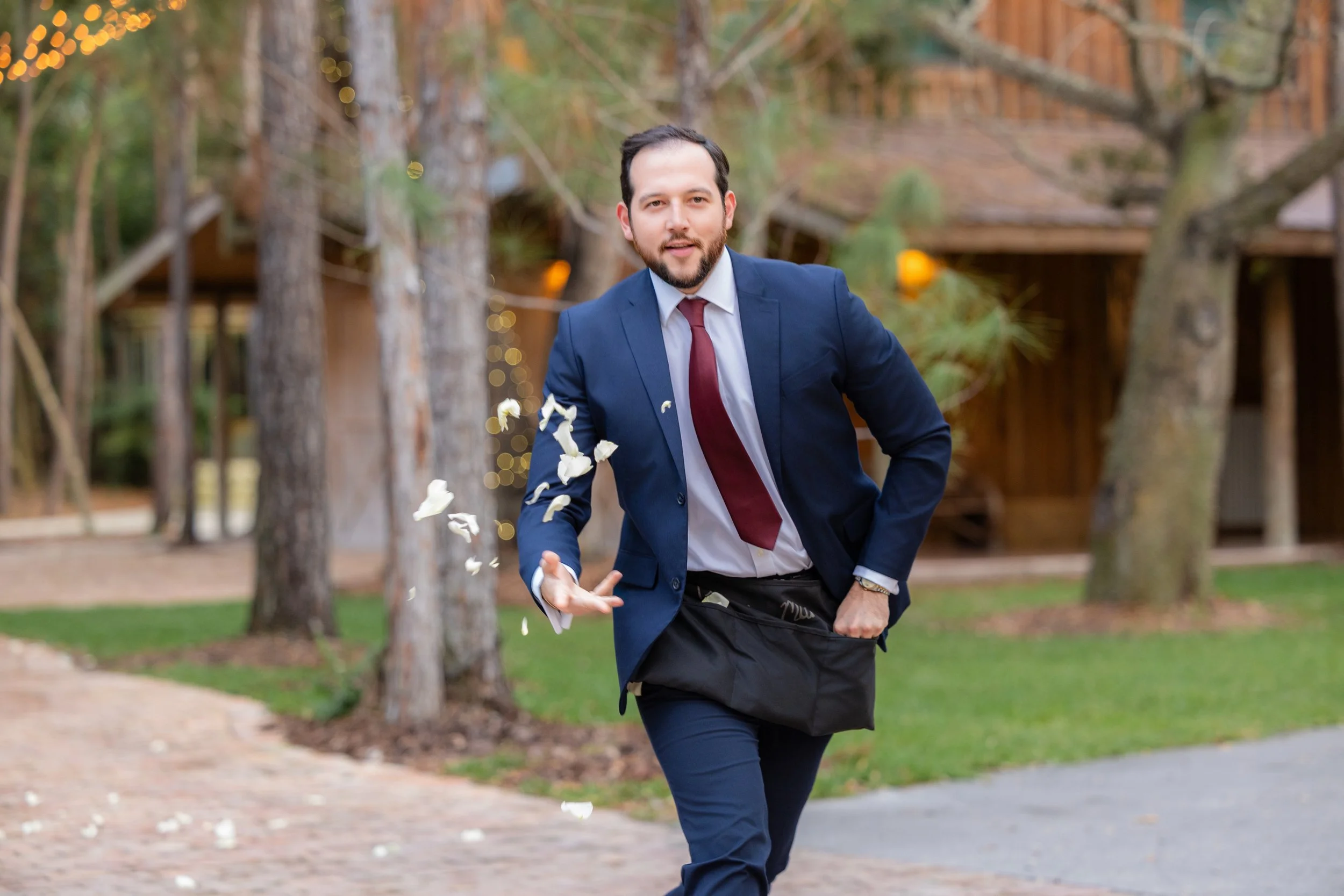 A man in a blue suit and red tie running outdoors with flower petals in motion, in a wooded area with trees and a wooden structure in the background.