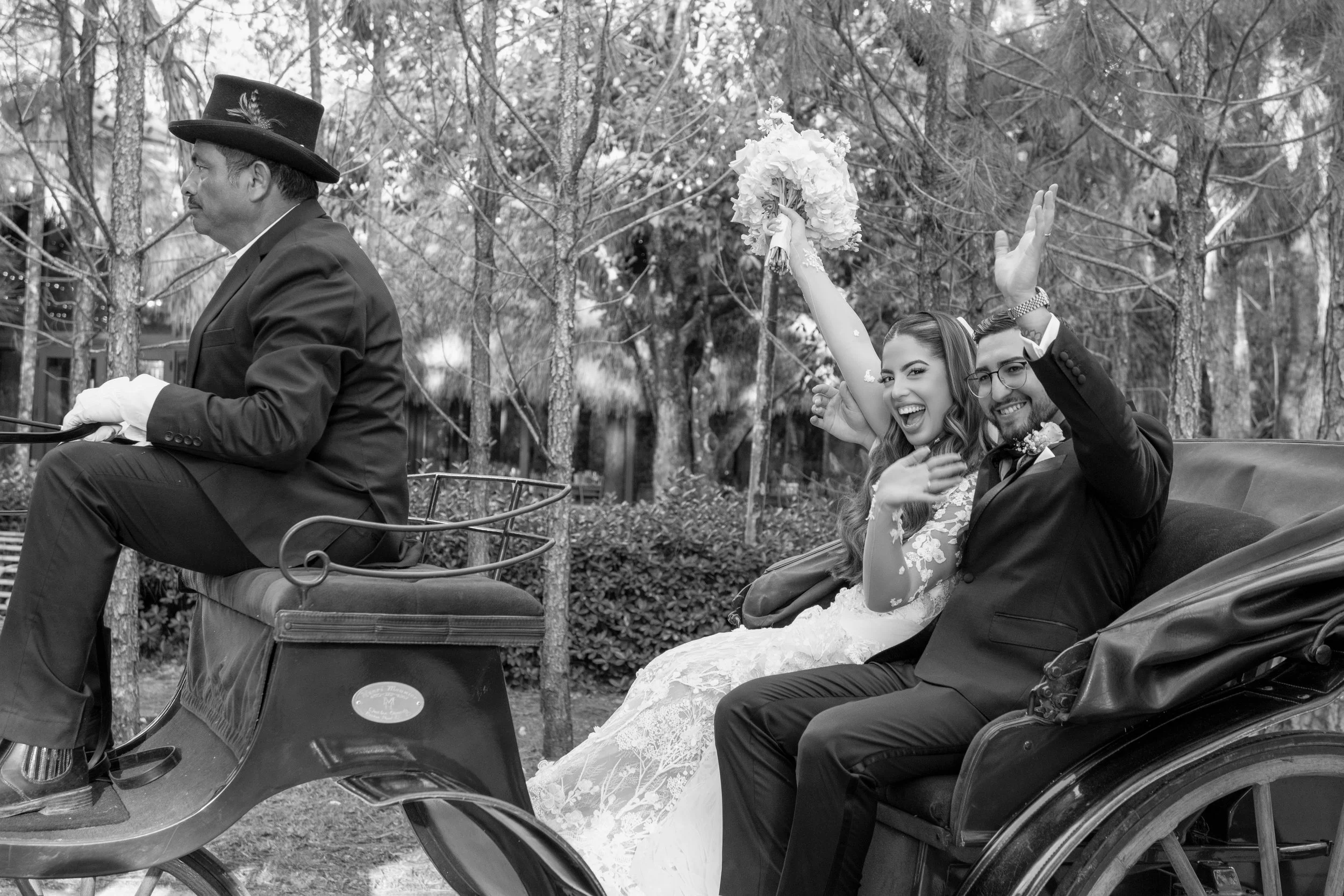 A bride and groom riding in a horse-drawn carriage, smiling and waving, with the bride holding a bouquet, outside in a wooded area.