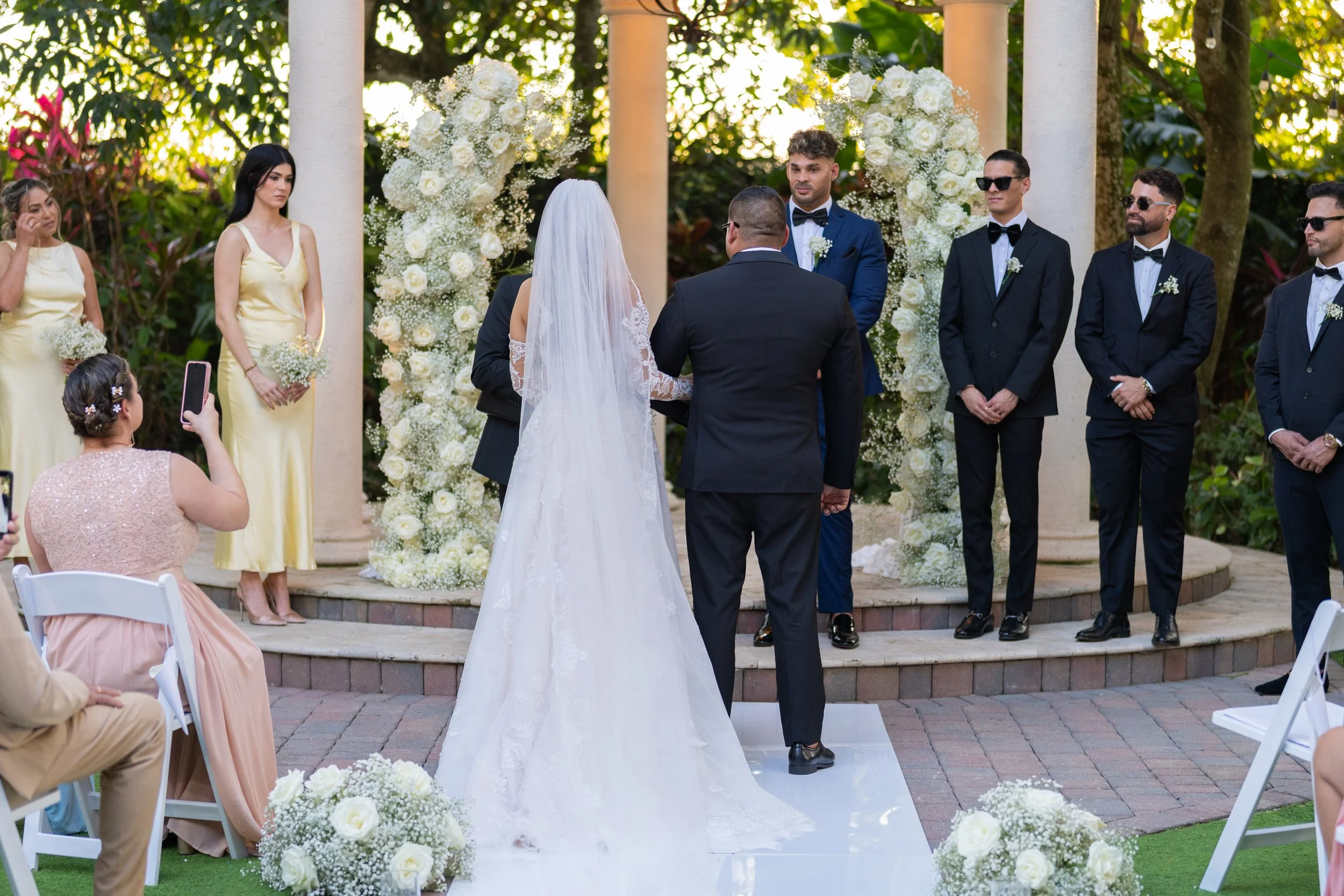 Wedding ceremony outdoors with the bride and groom facing officiant, surrounded by bridesmaids and groomsmen, with floral arches and guests seated, during daytime.