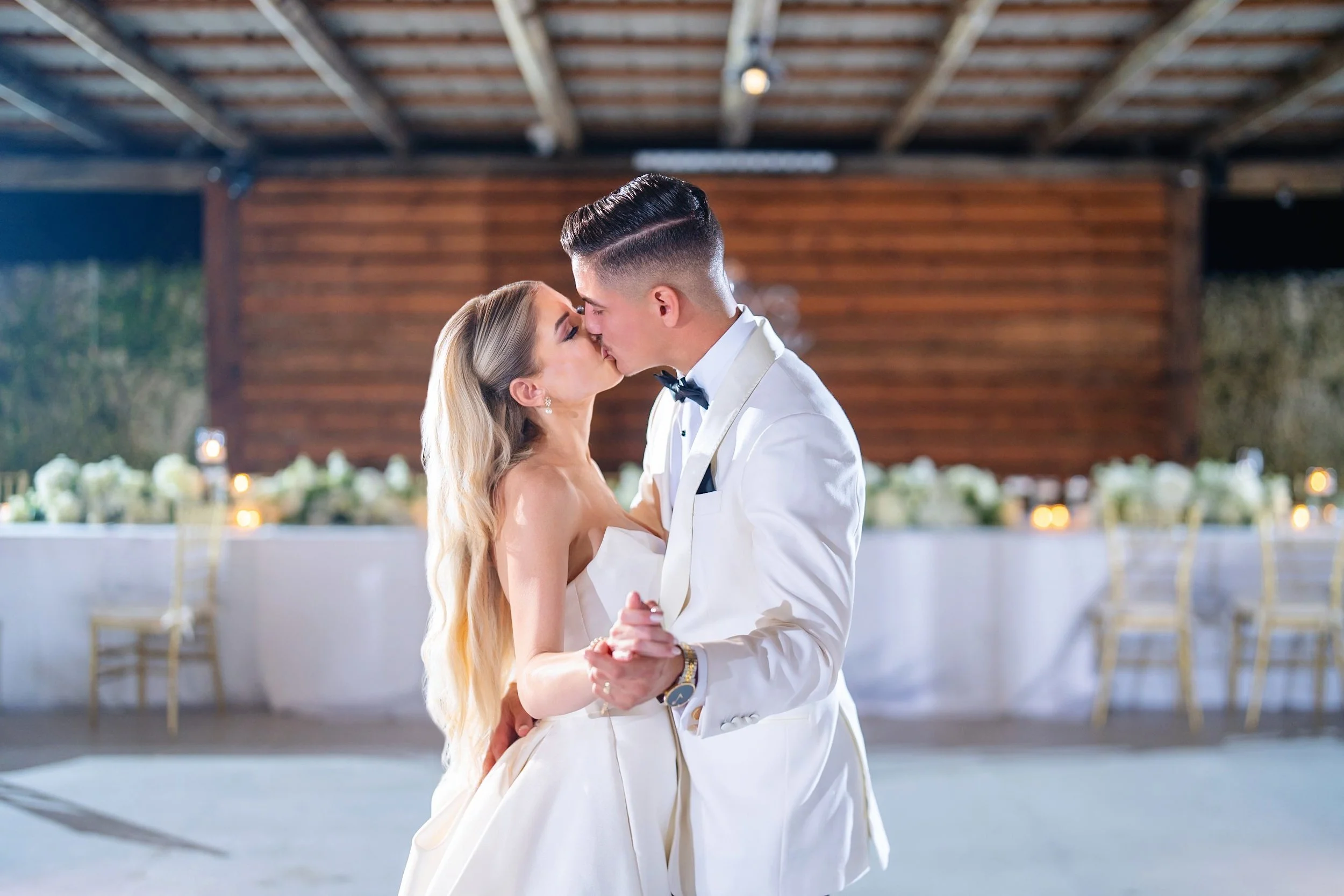 A bride and groom sharing a kiss while dancing at their wedding reception. The bride has long blonde hair and is wearing a strapless white gown, while the groom has dark hair and is dressed in a white tuxedo with a black bow tie. The background featu