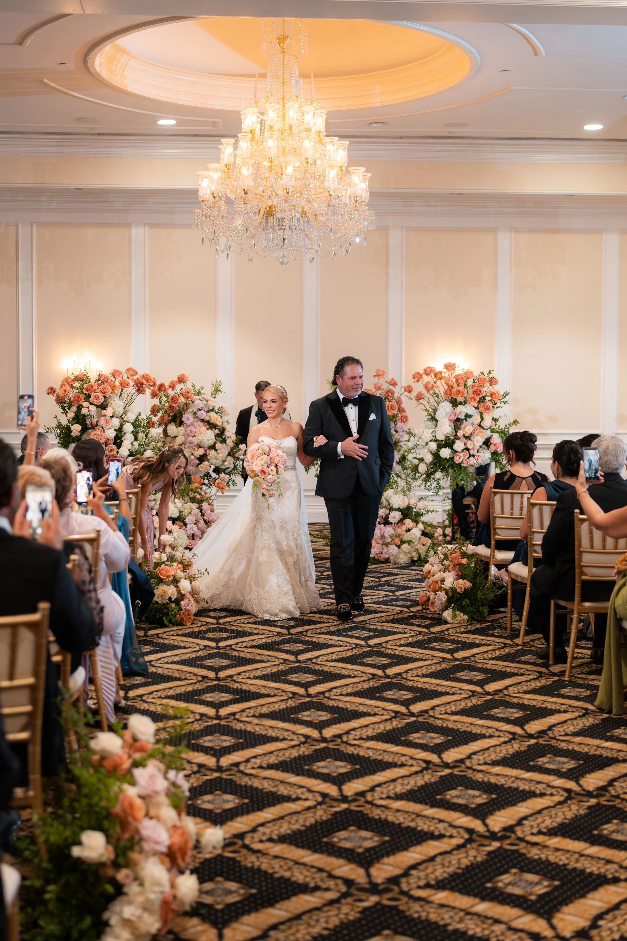 Bride and groom walking down the aisle at a wedding ceremony with guests taking photos, decorated with pink and white flowers and a large chandelier overhead.