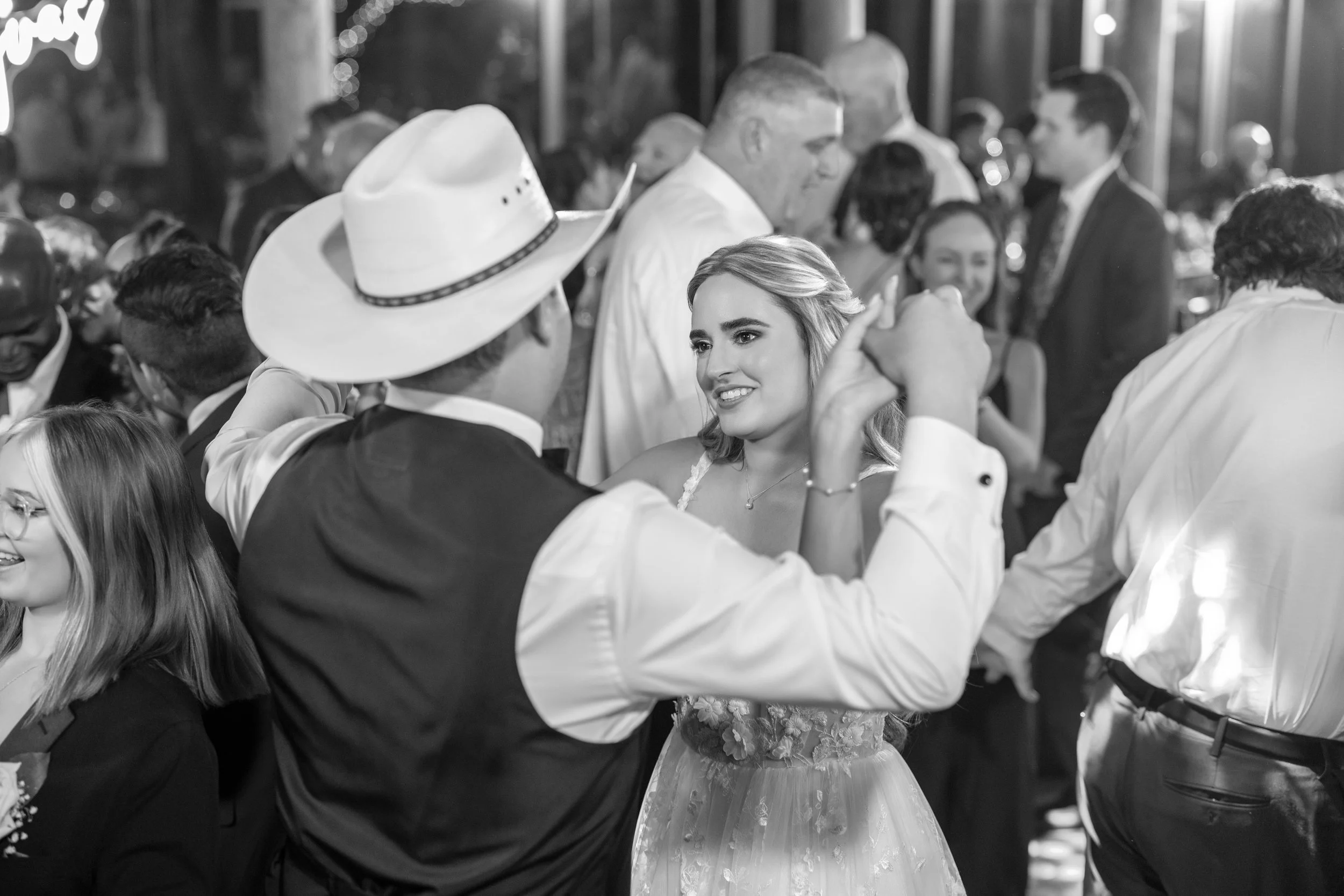 People dancing at a wedding reception, including a woman in a floral dress and a man in a cowboy hat.