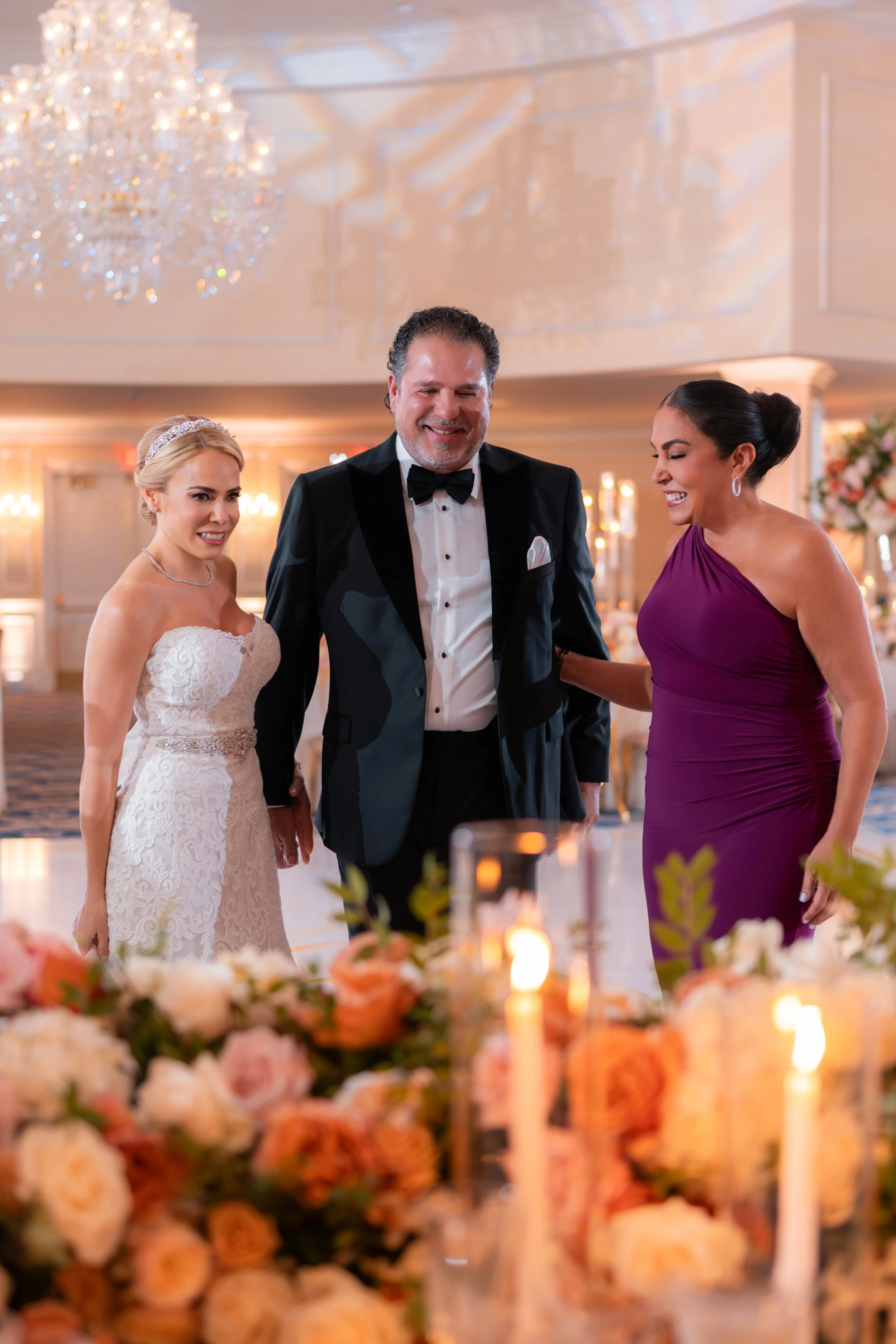 A bride in a white lace wedding gown, a groom in a black tuxedo, and a woman in a purple dress smiling together at a wedding reception.