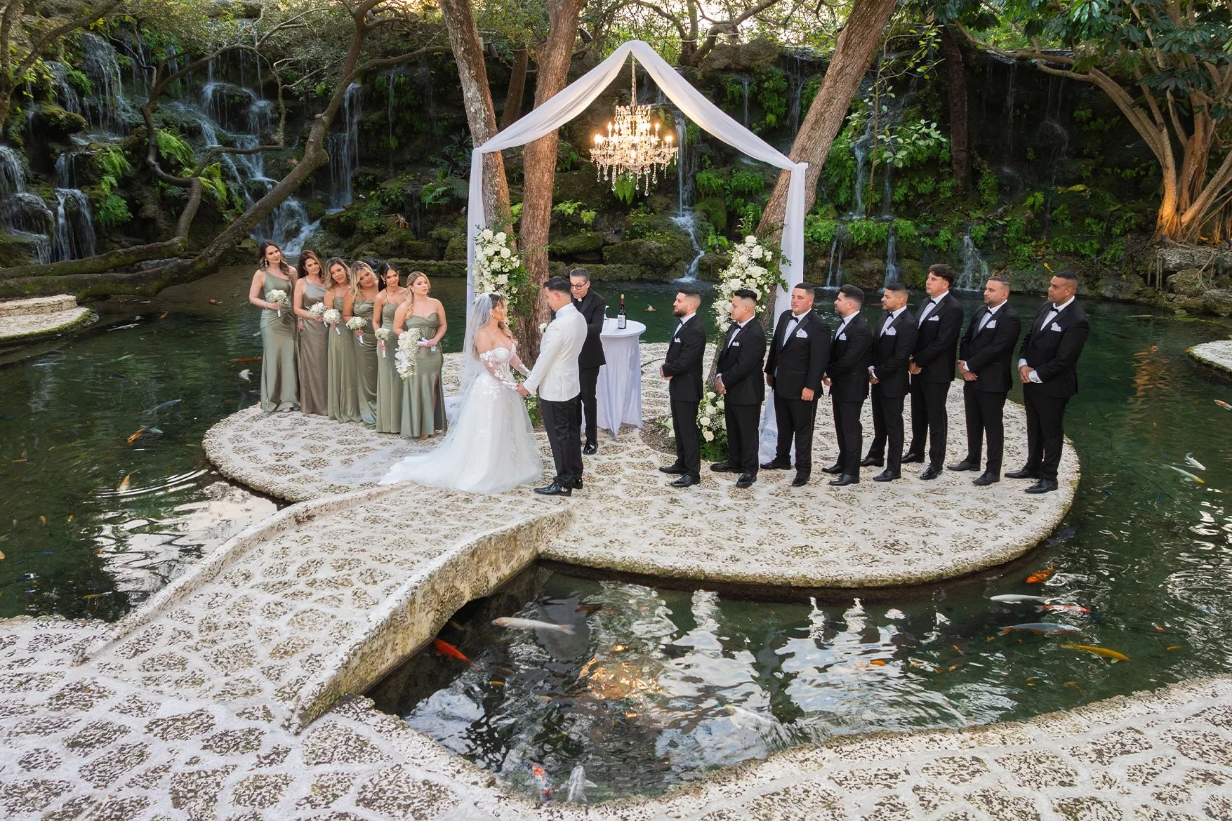 A wedding ceremony taking place on a small stone platform surrounded by water with koi fish. The bride in a white gown and veil is holding hands with the groom in a white suit. Bridesmaids in matching sage green dresses and groomsmen in black tuxedos stand on either side. An elegant chandelier hangs from a white canopy with floral arrangements, all set in a lush outdoor garden with a waterfall in the background.