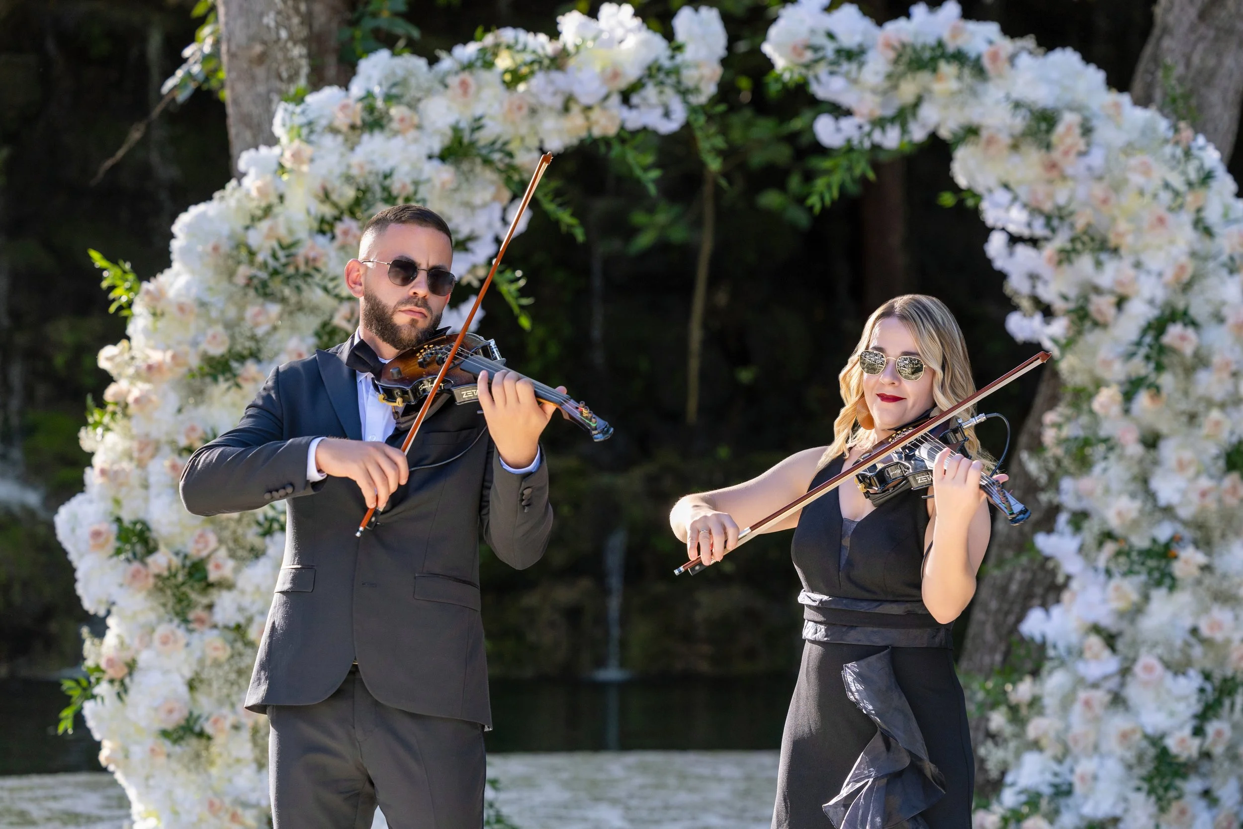 Two musicians, one man and one woman, playing violins outdoors in front of a floral arch with white and pink flowers