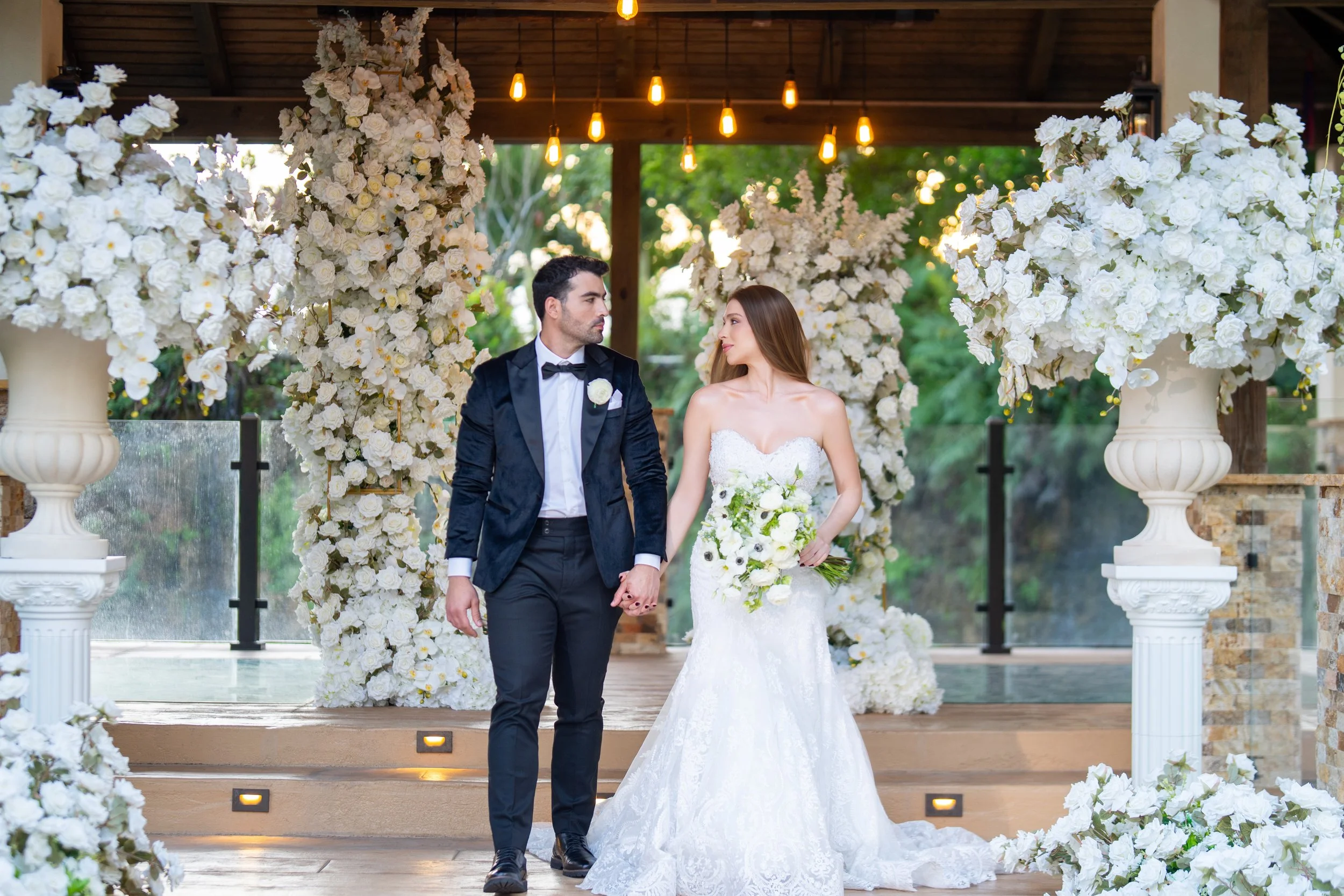 Bride and groom holding hands during their wedding ceremony, standing in front of a floral arch decorated with white flowers and lush greenery.