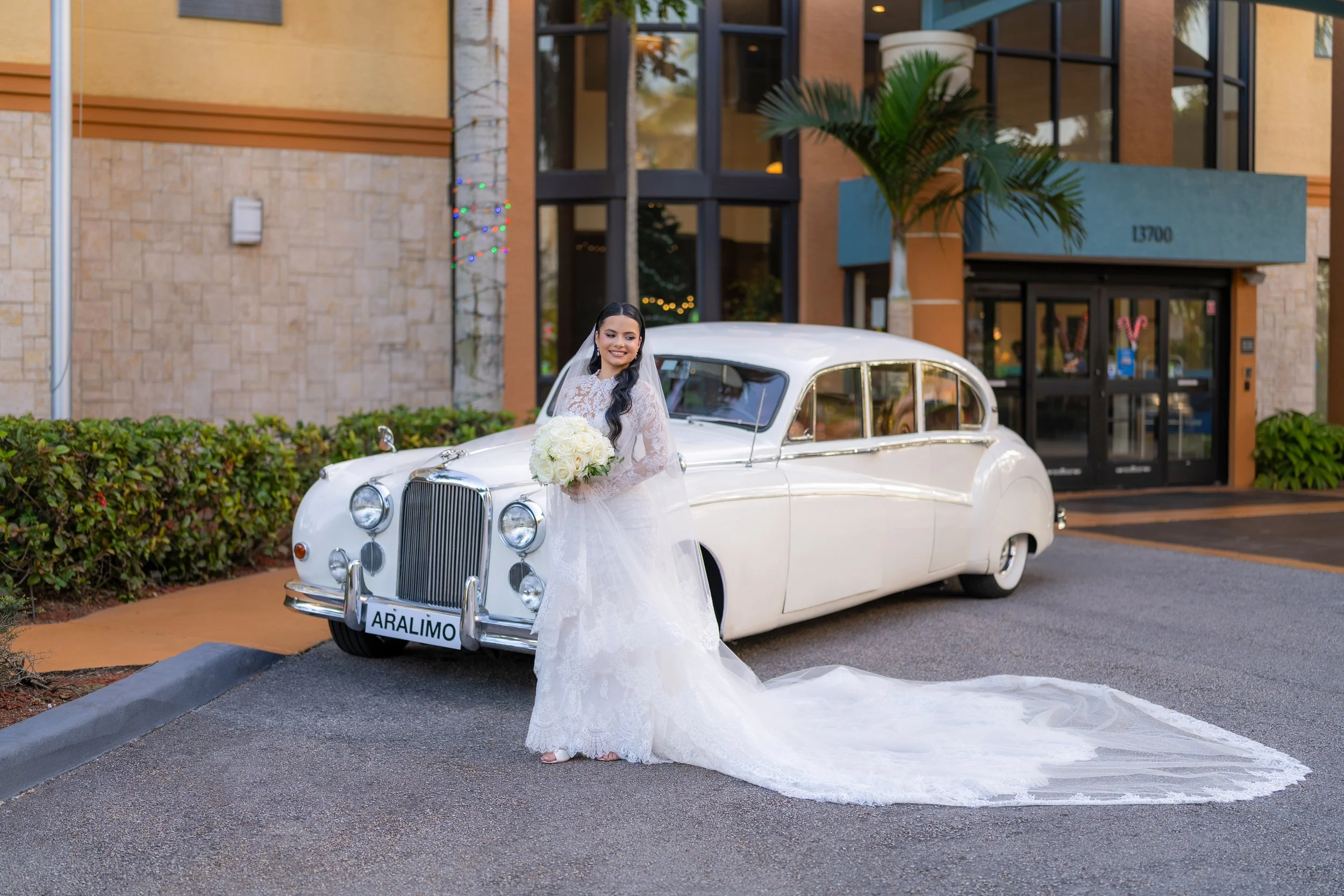 Bride in a white lace wedding dress holding a bouquet of white roses standing beside a vintage white car outside a modern building.