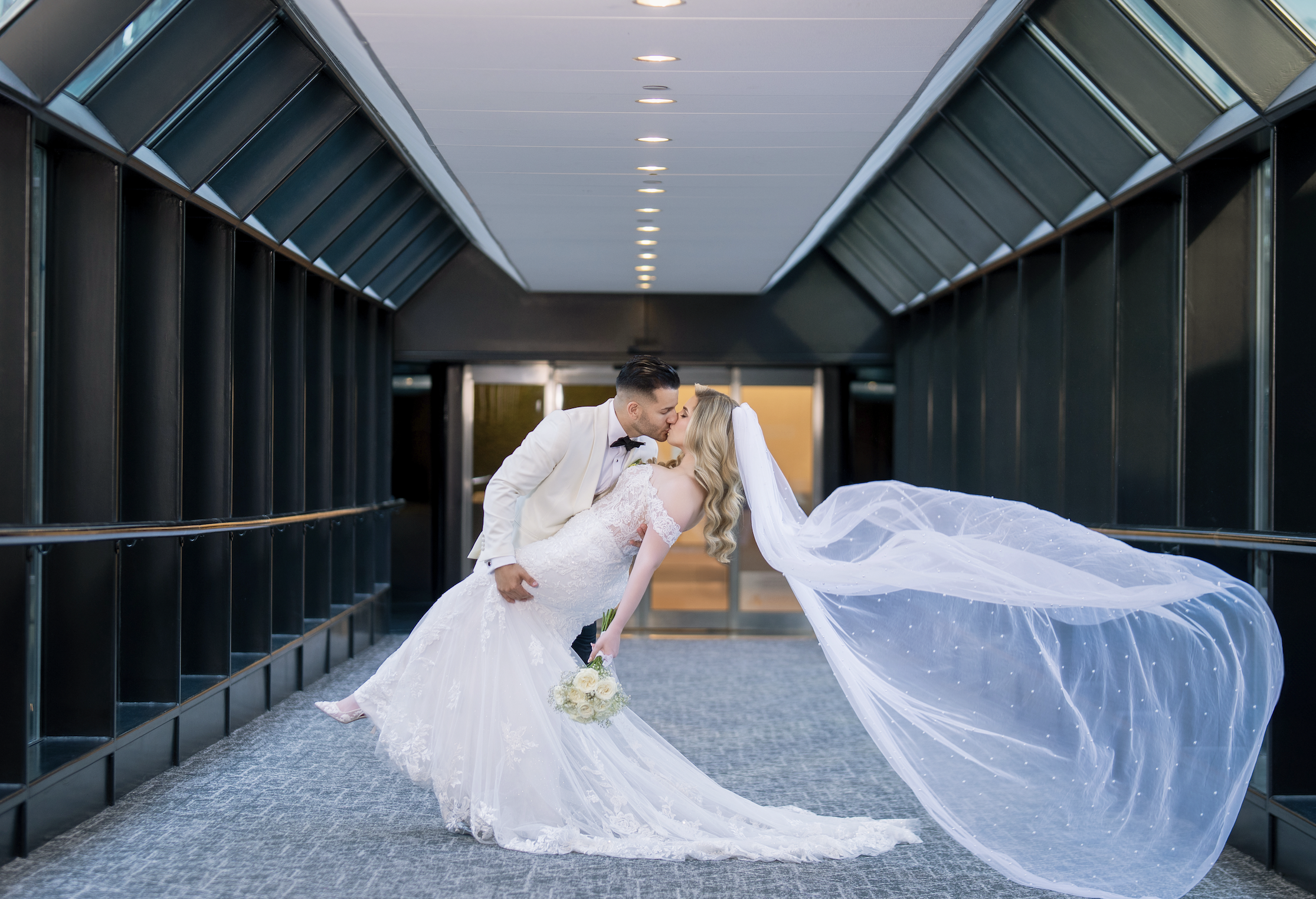 A bride and groom sharing a kiss in an indoor corridor, with the bride wearing a long white wedding gown and the groom in a white tuxedo with a black bow tie.