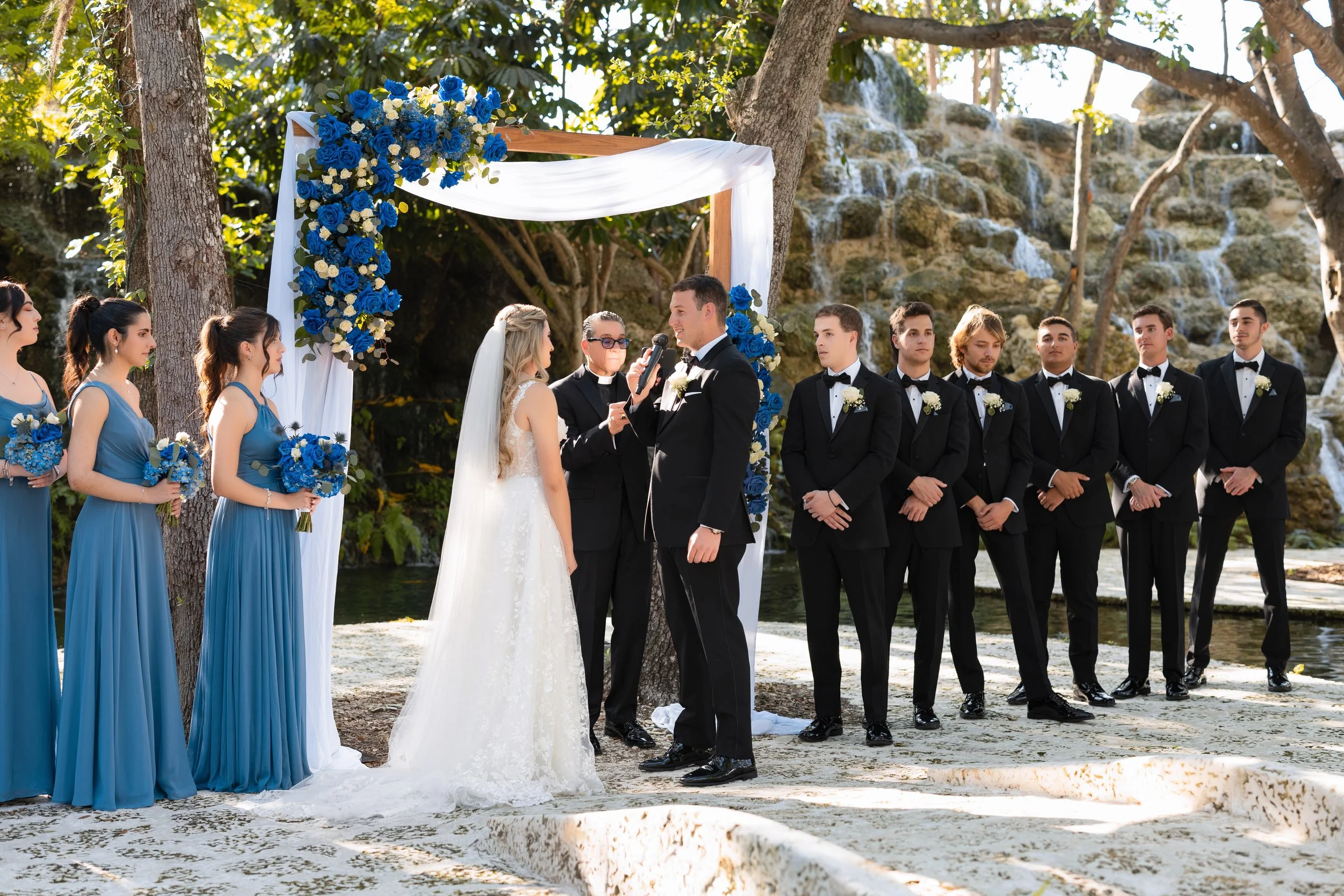 Wedding ceremony outdoors with a bride and groom exchanging vows under a decorated arch, surrounded by bridesmaids in blue dresses and groomsmen in black tuxedos, with trees and a waterfall in the background.
