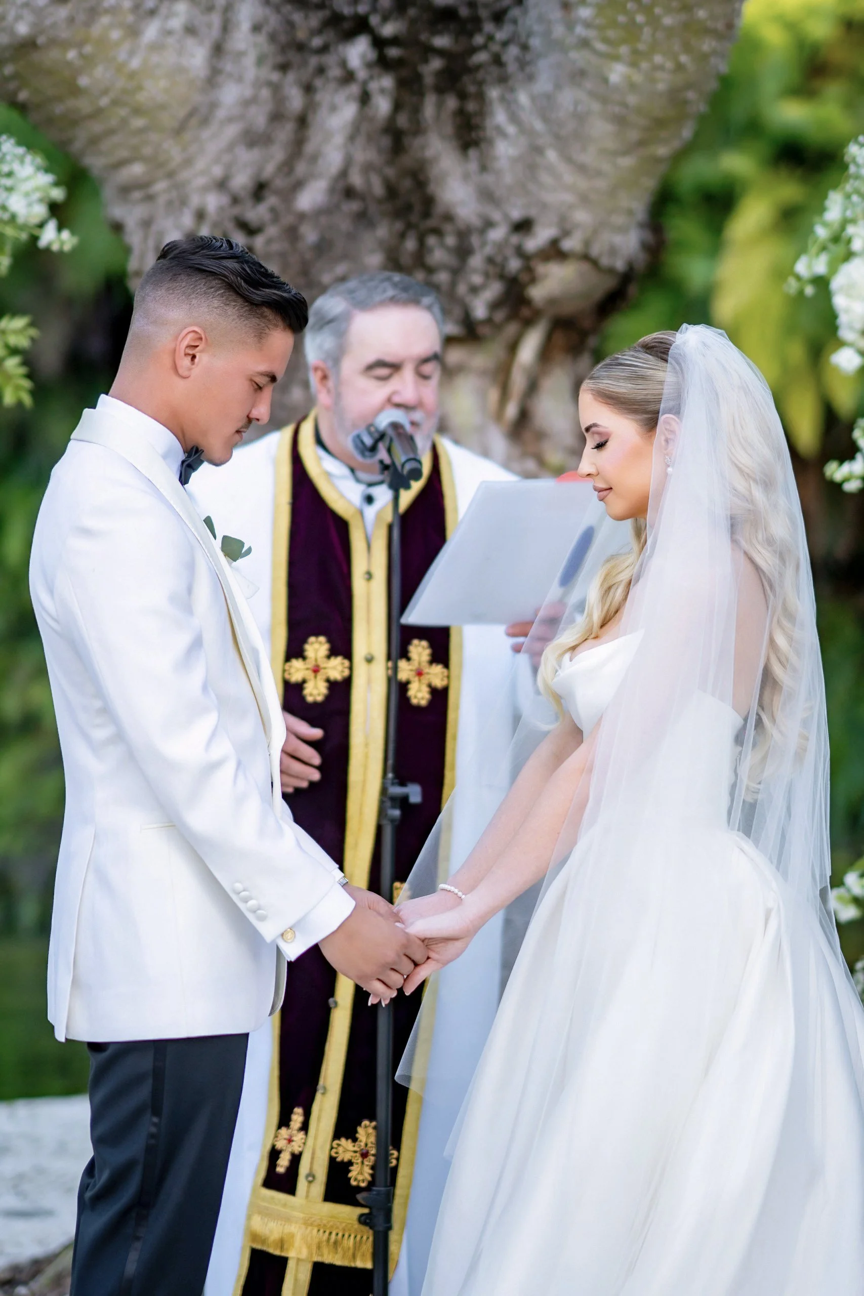 A couple getting married outdoors, holding hands and standing in front of an officiant reading vows, with greenery and a tree in the background.