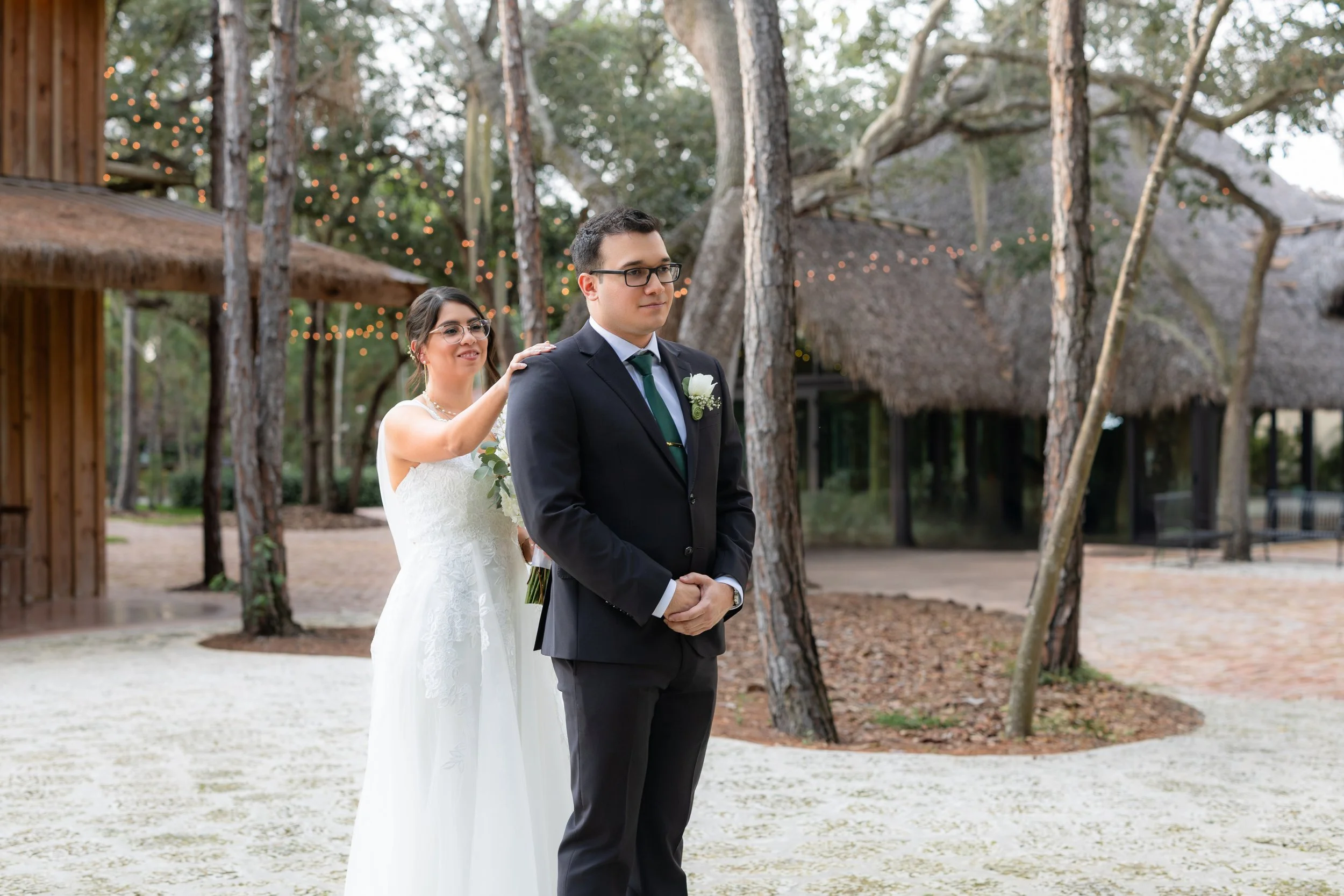 A bride standing behind a groom, gently placing her hand on his shoulder during a wedding ceremony outdoors. The scene takes place in a wooded area with fairy lights and a thatched-roof building in the background.