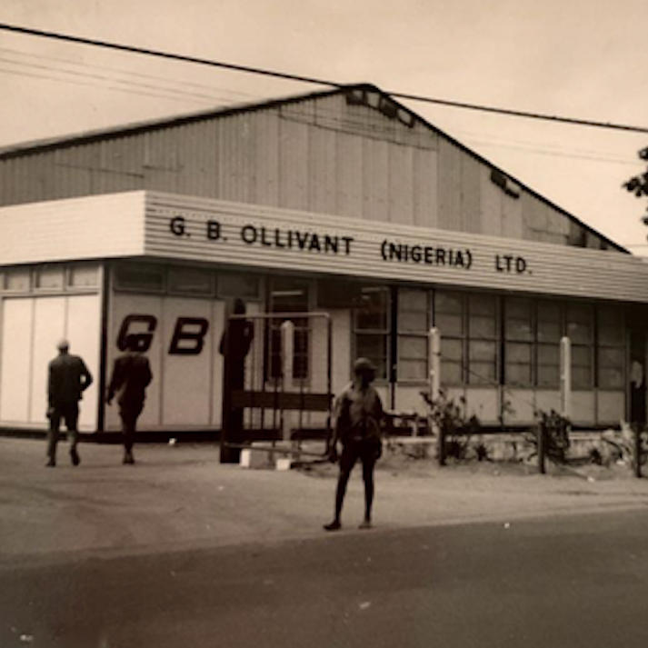 Historic black‑and‑white photo of the G. B. Ollivant Nigeria building with signage, warehouse façade and people walking outside.
