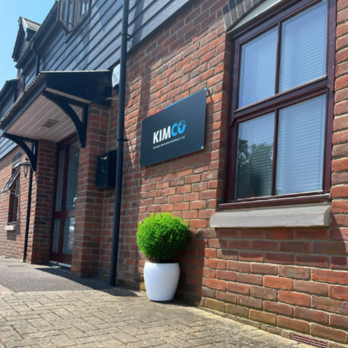 KIMCO headquarters with a branded exterior sign displaying “KIMCO” on a brick building, shown alongside a white planter and large office windows.