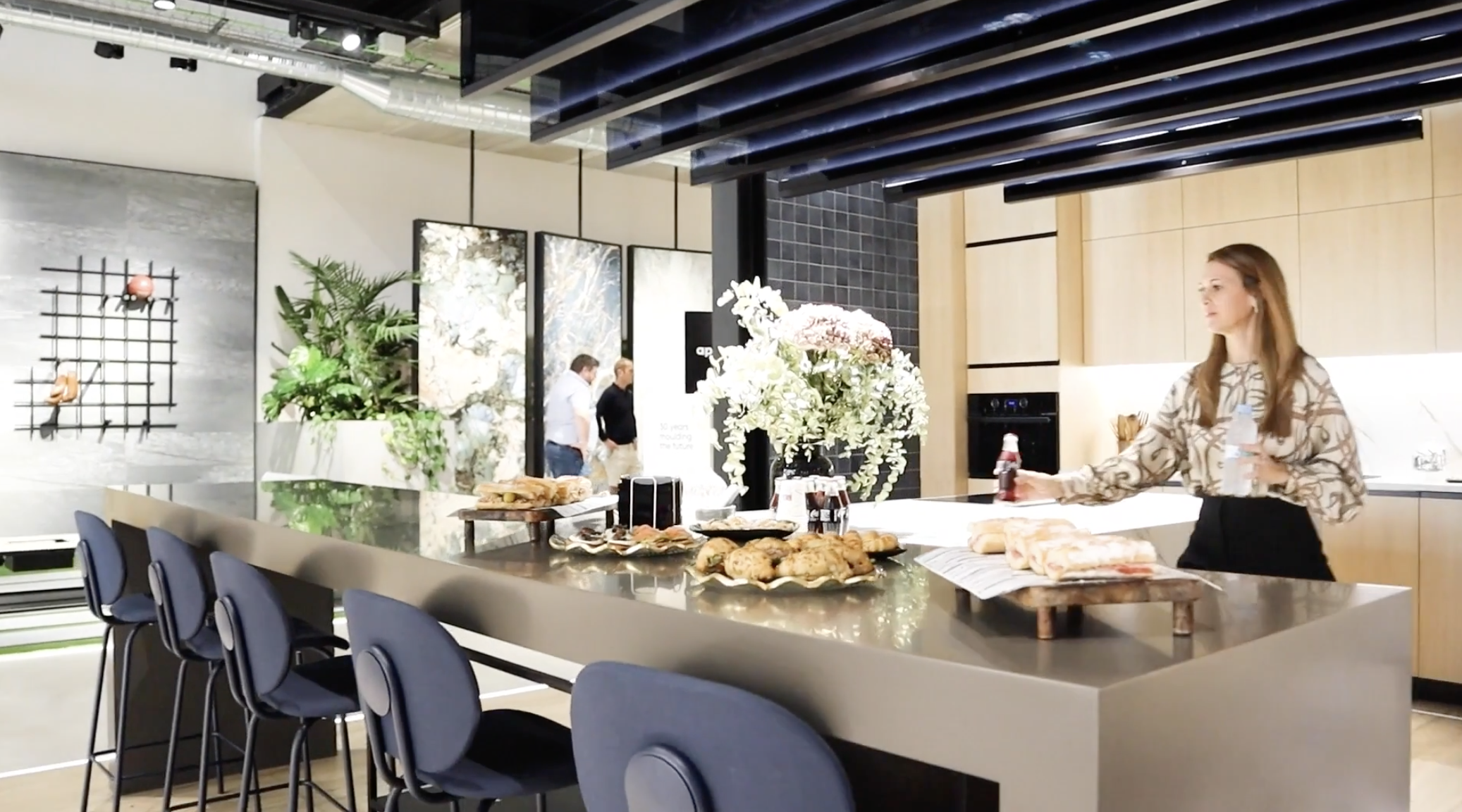 A modern kitchen or cafe interior with a large island counter displaying pastries, sandwiches, and drinks, adorned with a floral arrangement. A woman is reaching for a bottle, and three men are walking in the background.