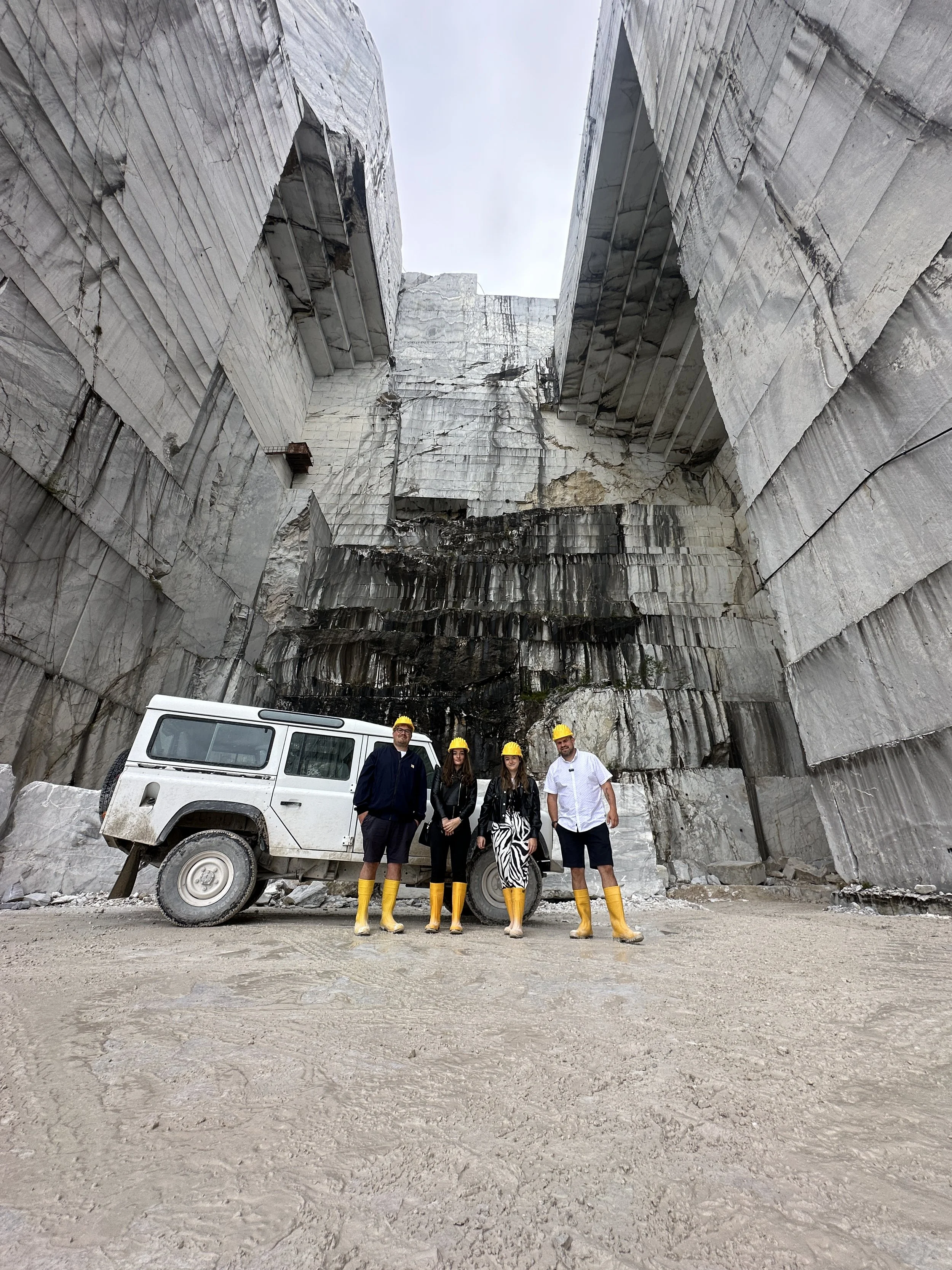 Four people wearing yellow safety helmets and yellow rubber boots standing in front of a large white off-road vehicle at a marble quarry. The quarry walls are high and rugged, with visible stratified layers of marble.
