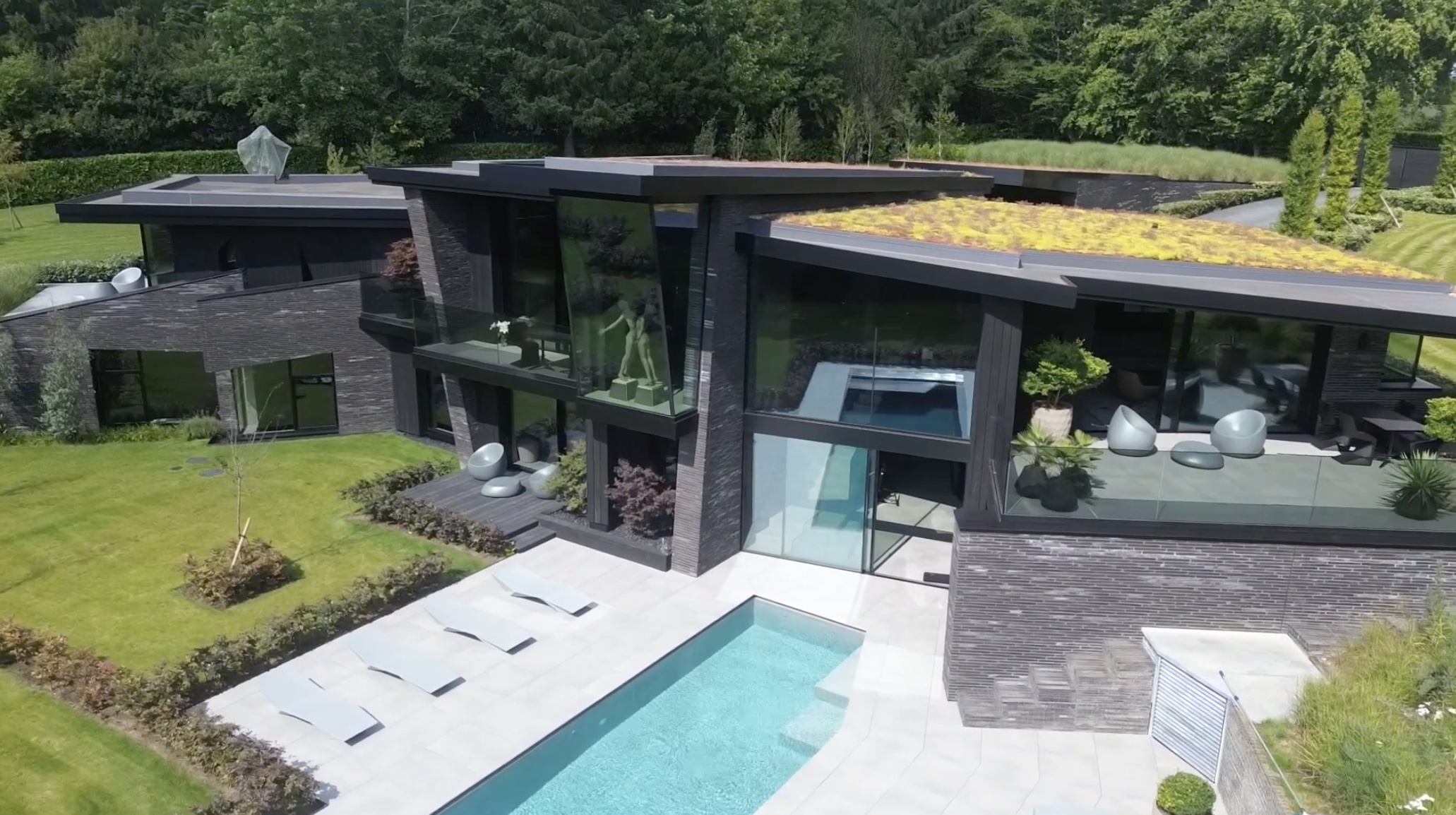 Modern house with large glass windows, a swimming pool, outdoor lounge area, green lawn, and lush greenery surrounding the property.