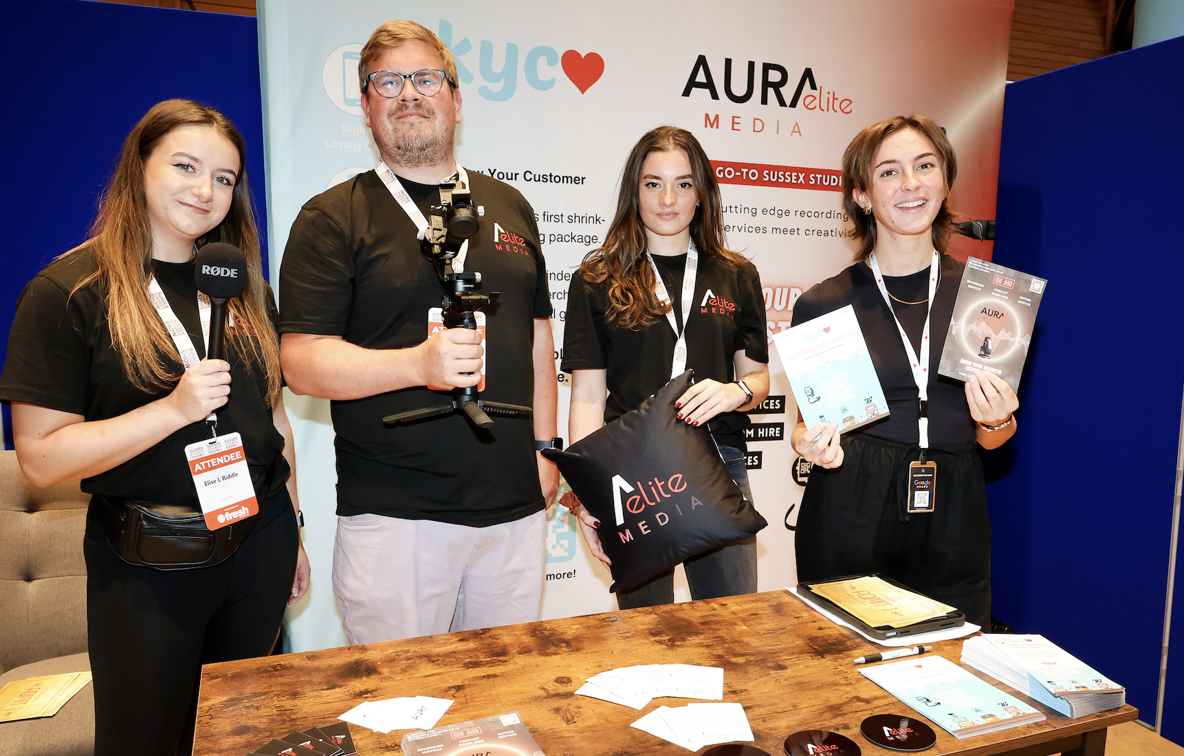 Group of four people at a trade show booth, holding branded items and promotional materials for Elite Media.
