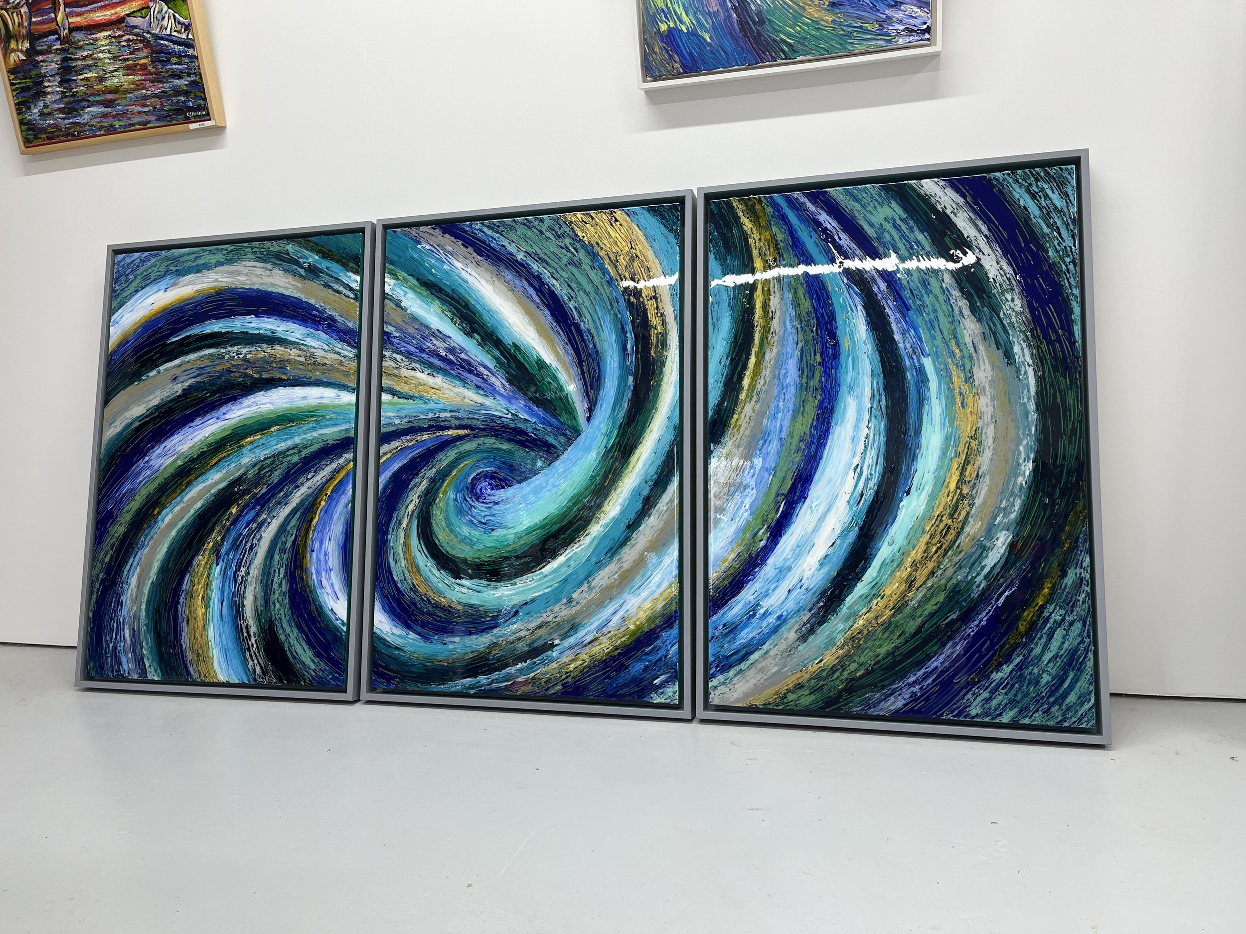 Three large modern abstract paintings featuring swirling patterns in shades of blue, green, white, and yellow, displayed on a white gallery wall.