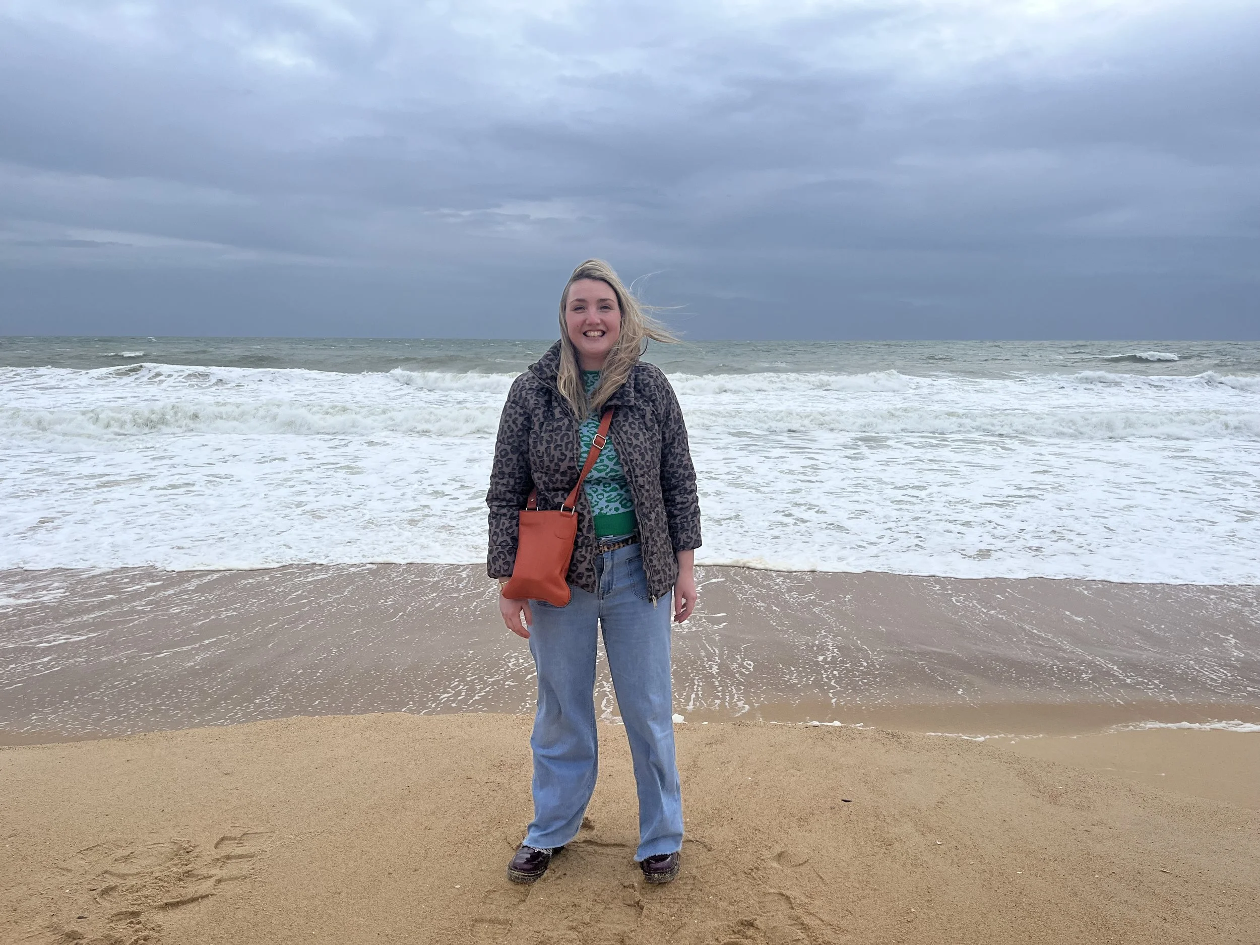 Chloe Pinheiro standing on the sandy beach with ocean waves behind her, under a cloudy sky.
