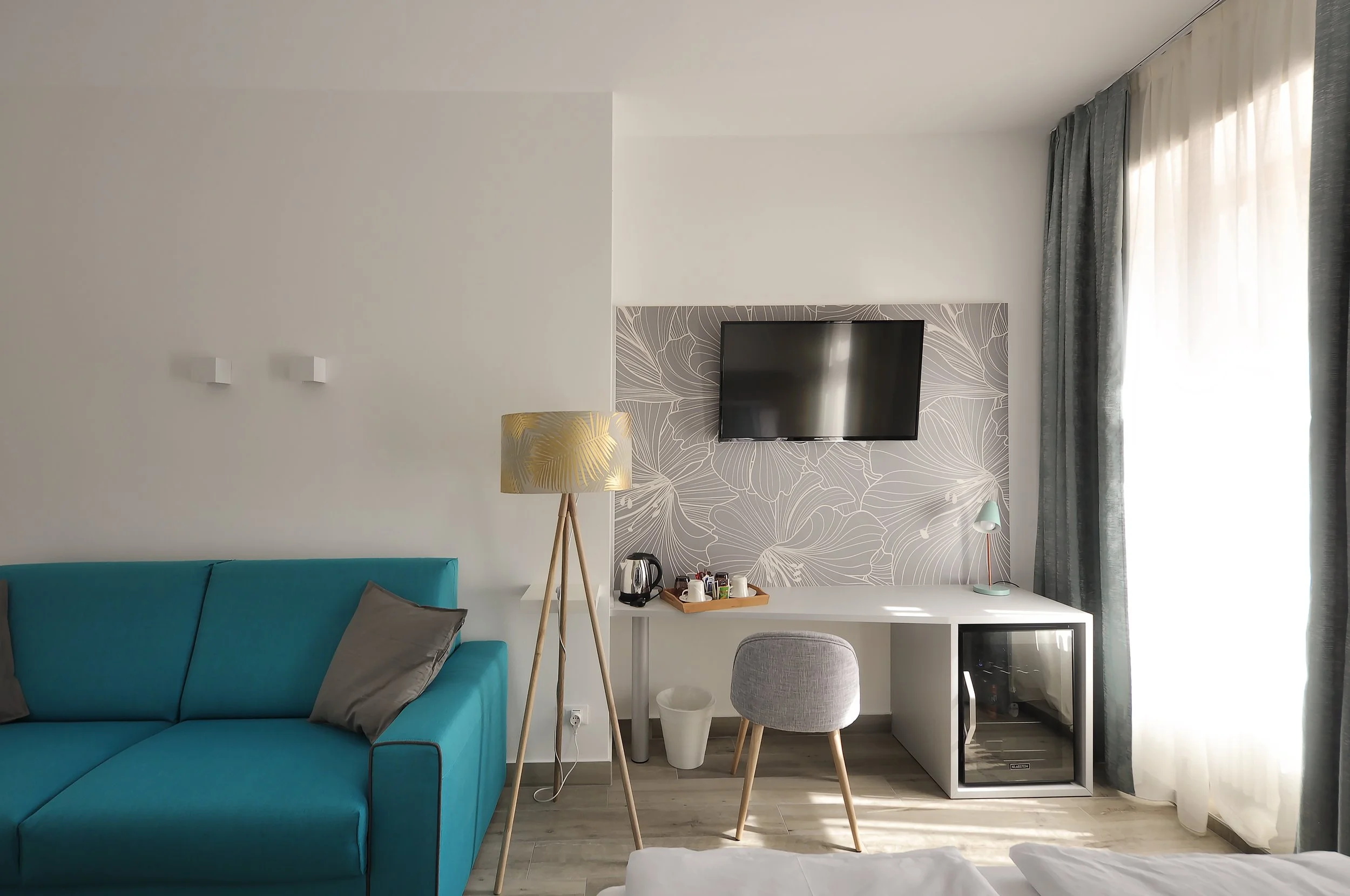Interno di una stanza d'albergo con divano azzurro, lampada da terra, scrivania bianca con sedia grigia, televisore a parete, vetrinetta refrigerata, e tende verdi a destra, illuminata dalla luce naturale.