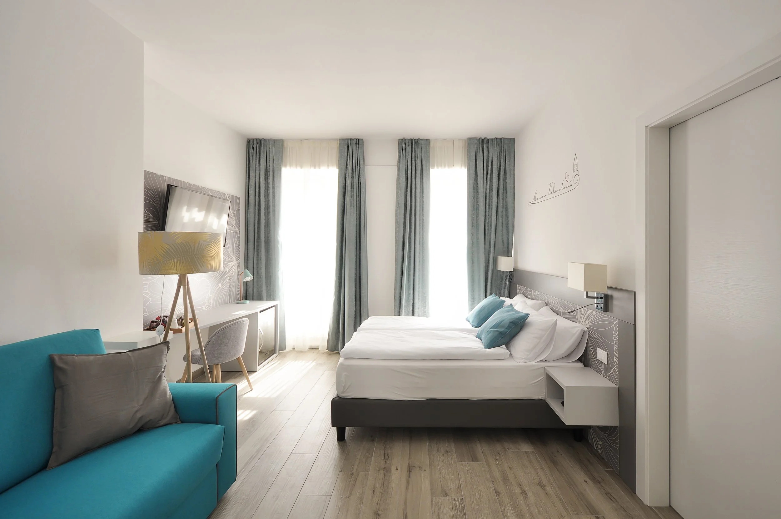 Camera moderna con letto matrimoniale, grande finestra con tende grigie, divano blu, scrivania e lampada da terra.