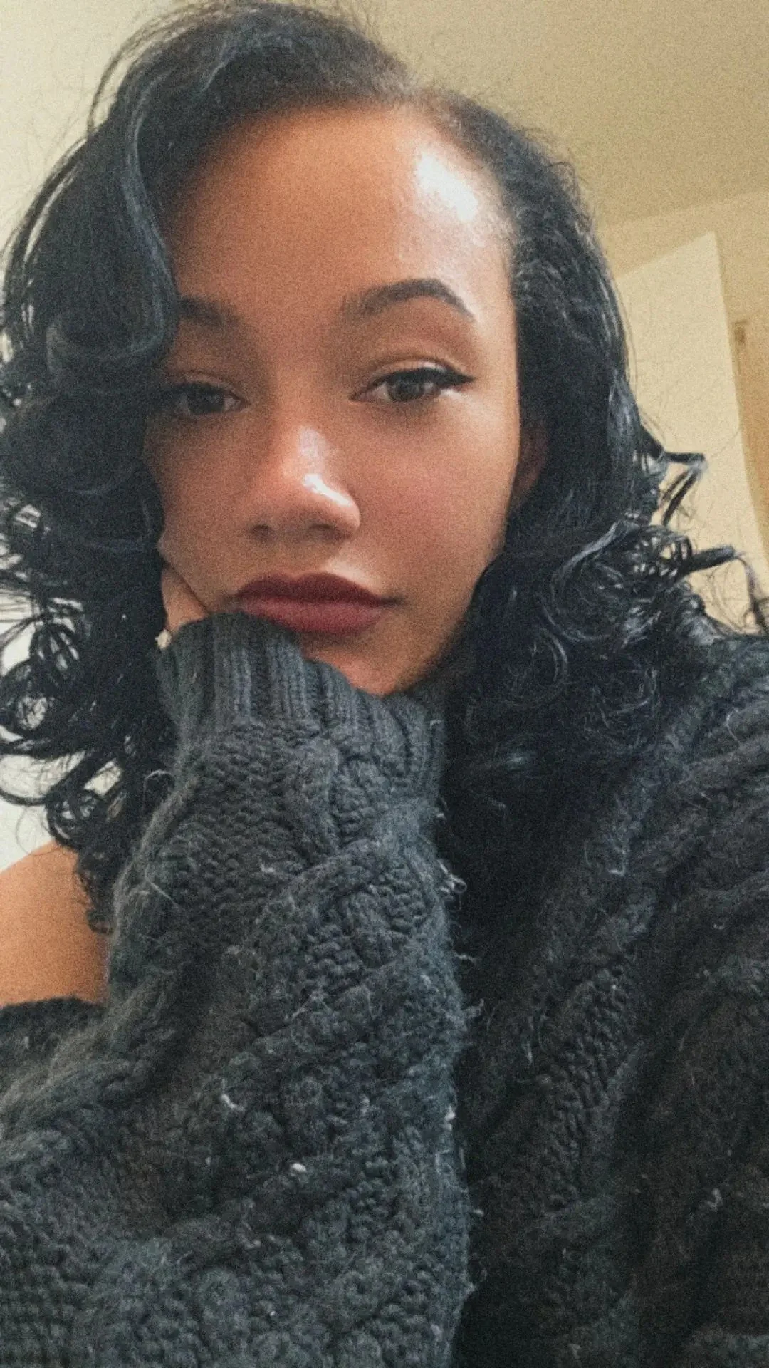 A young woman with curly black hair and red lipstick resting her chin on her hand, wearing a dark sweater.
