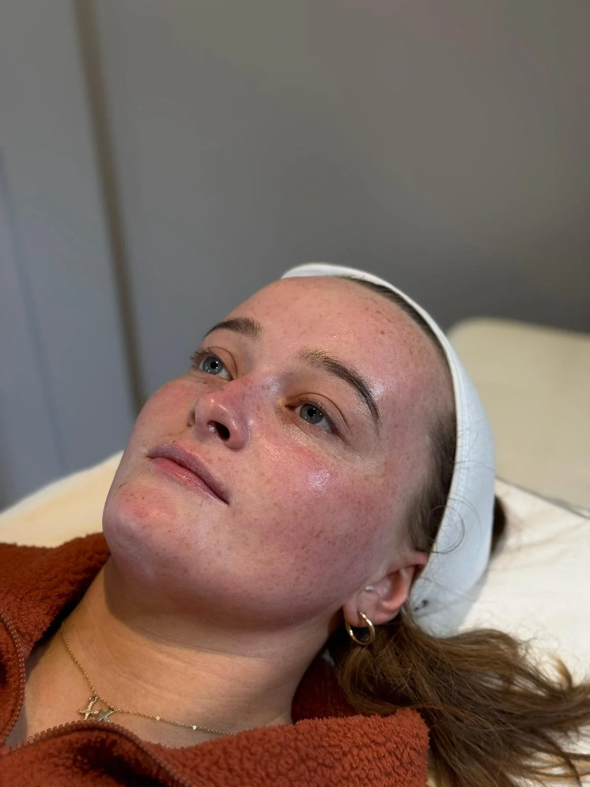 Close-up of a woman with clear skin and freckles lying on a pillow, wearing gold hoop earrings and a gold necklace, with her head slightly tilted, indoors.